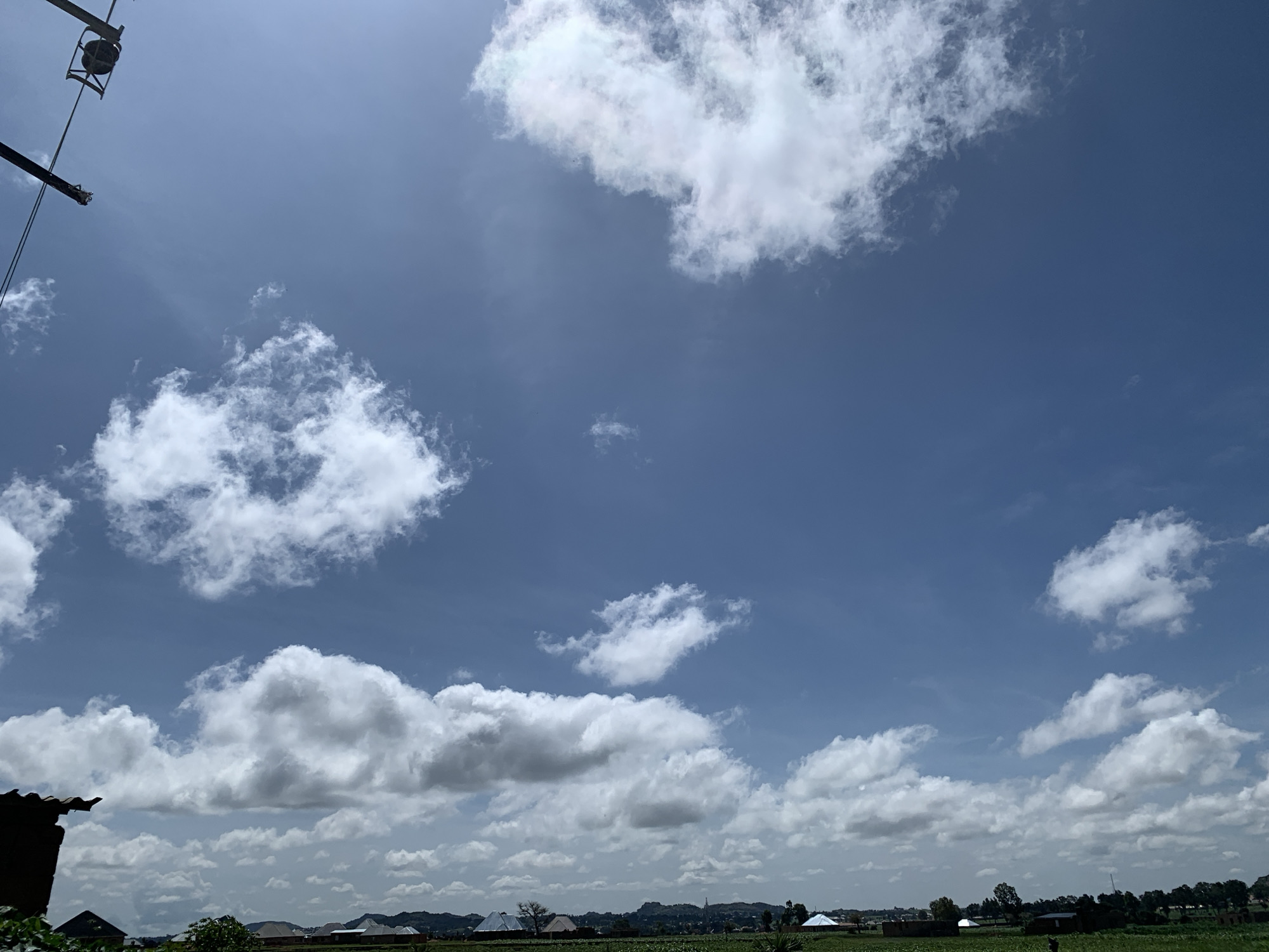 The image depicts a bright, sunny day in Kuru Karama, Nigeria, characterized by a vast expanse of blue sky dotted with fluffy white clouds. In the foreground, on the left side of the image, a dark, angular structure, possibly a roof or part of a building, is visible. A dark pole, likely a utility pole, extends from the top left corner, with wires connected to it. The middle ground reveals a landscape of scattered buildings with varied roofing, suggesting a semi-urban or rural setting. Green fields and trees are interspersed throughout the landscape, indicating agricultural activity or natural vegetation. The distant background is marked by rolling hills and more scattered structures, contributing to the overall impression of a sun-drenched environment under a dynamic sky.