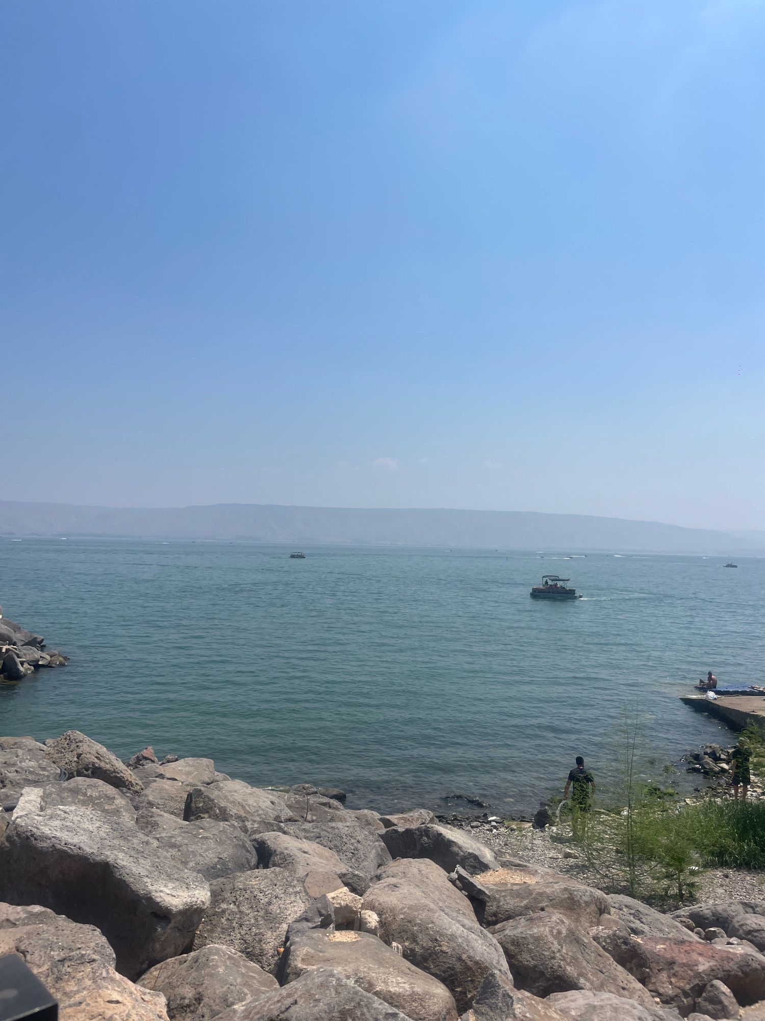 This is a wide-angle, eye-level shot of the Sea of Galilee in Tiberias, Israel, on a clear, hazy day. In the foreground, large, grey rocks line the shore, with the tranquil blue water of the sea stretching out to the horizon. Several boats, including a pontoon boat with people on it, are visible on the water. In the mid-ground, a low, hazy mountain range forms the distant shoreline. On the right side of the image, a concrete pier extends into the water, where a person is seated. To the left of the pier, a person in a dark shirt is standing near the water's edge. The sky is a pale, clear blue.