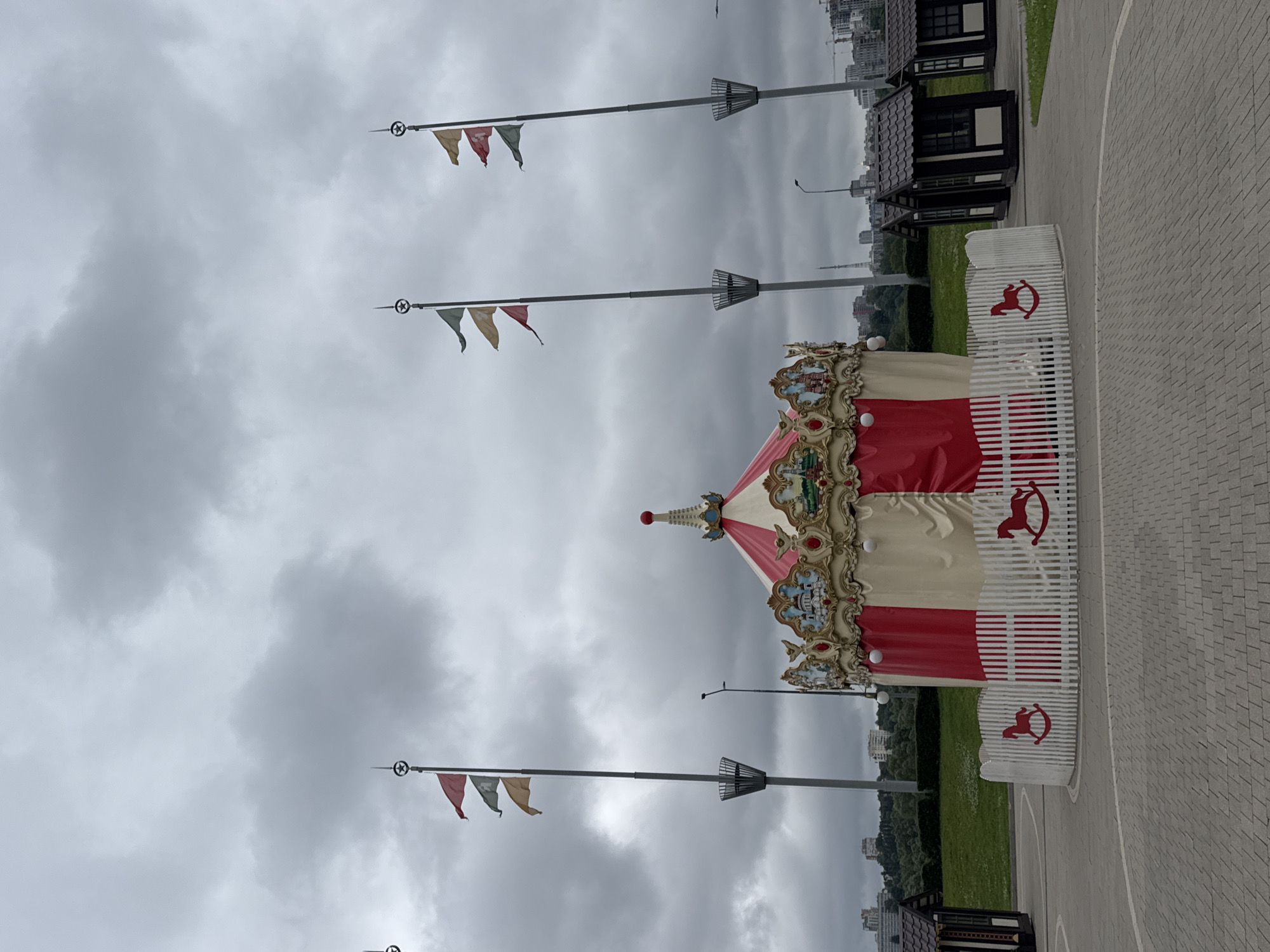 The image depicts an outdoor scene in Nizhny Novgorod, Russia, featuring a carousel and several small structures. The sky is overcast with dark, heavy clouds, suggesting a gloomy or stormy day.
In the foreground, a red and white striped carousel with ornate gold trim is prominently displayed. The carousel is fenced with a white picket fence that has silhouettes of rocking horses painted in red. To the left and right of the carousel are small, dark-colored wooden kiosks with tiled roofs, resembling miniature houses or booths. One kiosk is visible on the far left, and two are partially visible on the right.
Behind the carousel, a grassy slope is visible, leading up to a line of trees. In the distance, the cityscape of Nizhny Novgorod can be seen, with buildings and a telecommunications tower under the cloudy sky.
Several tall poles with flags are positioned behind the carousel and to the sides. The flags are of various colors, including red, green, and yellow, and are attached to poles topped with a star-shaped finial. The overall impression is that of an amusement area or a park setting experiencing inclement weather. No people are visible in the image.