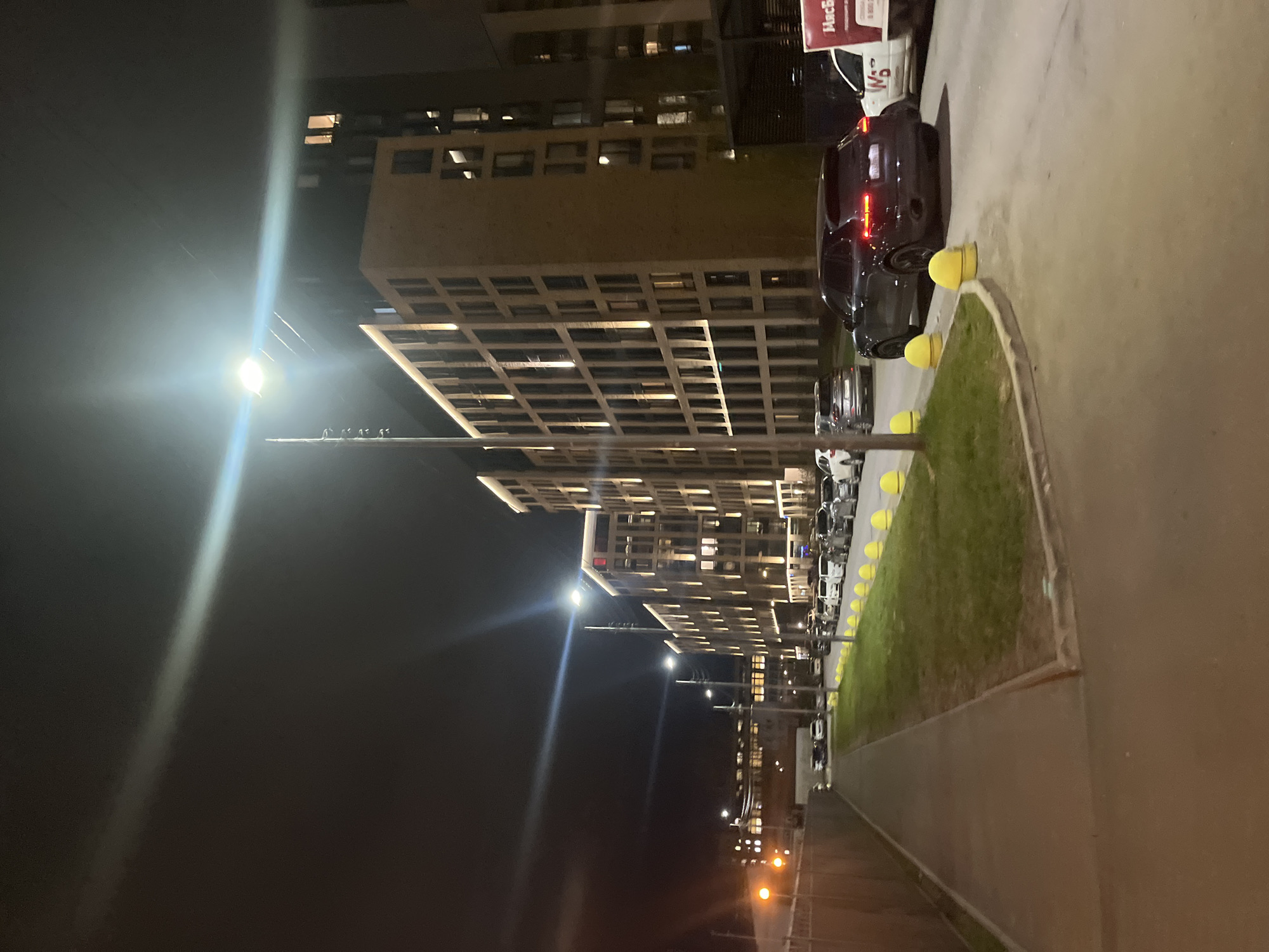 This image captures a nighttime urban scene in Saint Petersburg, Russia. The dominant subjects are modern, multi-story apartment buildings with numerous windows, many of which are illuminated, suggesting occupancy. These buildings feature a distinctive grid-like facade.
On the right side of the image, a paved road or parking area is visible, with several cars parked in a line. A dark-colored car with its red taillights illuminated is prominent in the foreground. Adjacent to the road is a strip of green grass, neatly bordered by a series of bright yellow, spherical bollards. Beyond the grass strip, another paved area, likely a sidewalk or pedestrian path, extends into the distance.
The scene is brightly lit by multiple streetlights, which create significant lens flares in the dark night sky. No people are visible, and no specific activity or event is taking place, presenting a quiet, static view of an urban environment after dark. The weather appears clear.
In the upper right corner, a red and white sign is partially visible, displaying the Cyrillic text "МаксБел" and "W5", along with a partially visible phone number, "8 800 234...". The overall impression is of a contemporary, well-lit urban area during the evening hours.
