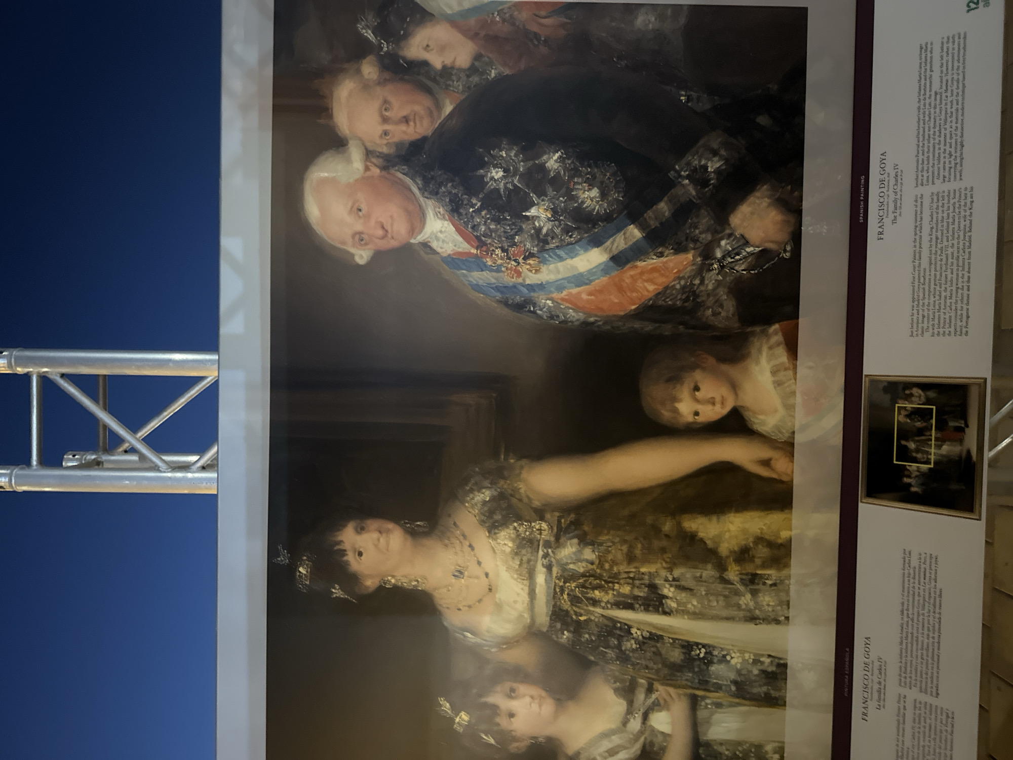 The image shows a framed reproduction of Francisco de Goya's painting "The Family of Charles IV." The painting depicts the Spanish royal family, including King Charles IV, his wife Maria Luisa of Parma, and their children and other relatives. The reproduction is displayed outdoors, with a blue sky and a metal structure visible in the background, suggesting it might be part of an exhibition or public display. Text panels with information about the painting and the artist are visible below the reproduction. The location is identified as Mazarrón, Spain, where a sign indicates "PINTURA ESPAÑOLA" (Spanish Painting). The reproduction is large, with a prominent gold frame. The painting itself is detailed, with the figures in formal attire, and Goya's characteristic use of light and shadow is evident.