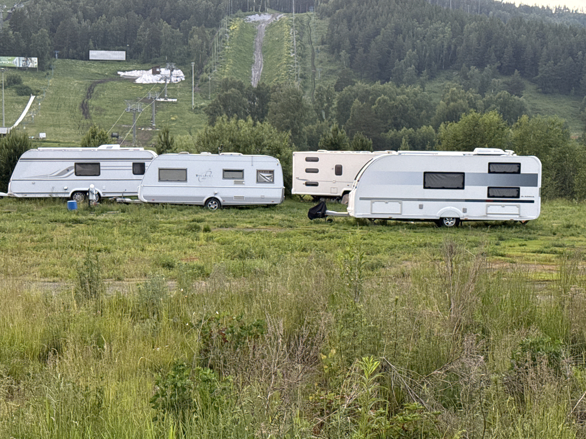 The image depicts a rural scene in Syrostan, Russia, featuring several parked caravans in a grassy field. In the background, a wooded hill is visible, with a ski slope marked by a dirt track and ski lift pylons. The ski slope appears to have a patch of snow at its base, suggesting it is either the end of the season or a facility for snowmaking. Two white caravans are prominent in the foreground, parked side-by-side. To the right of these, a cream-colored caravan is also visible, with a black cover over its hitch. The environment is characterized by tall, uncut grass and scattered vegetation. The overall impression is of a quiet, undeveloped area, likely used for recreation or camping.