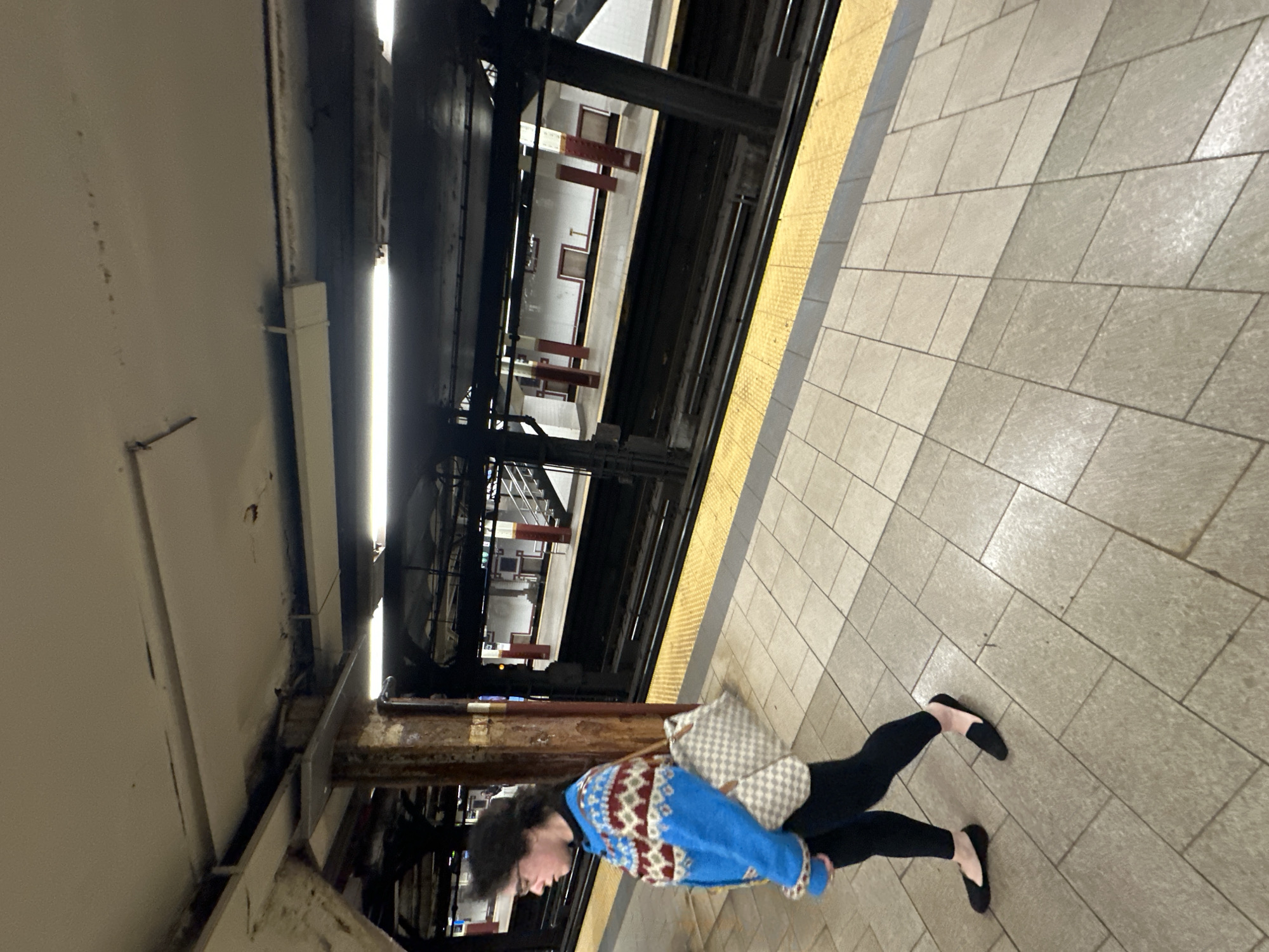 The image captures an underground subway platform scene, presented from a slightly elevated perspective. In the foreground, a young person stands on the light-colored, textured tiled platform. They have dark, curly hair and wear glasses, a distinctive blue sweater with a red and white geometric pattern, black pants, and black flat shoes. A checkered grey and white shoulder bag hangs from their left shoulder. Their head is turned to the left.
The platform they are on features a prominent yellow tactile warning strip running along its edge, separating it from the dark subway tracks. Across these tracks, another subway platform is visible, featuring similar light-colored walls and what appear to be benches. Above the tracks and between the two platforms, a complex network of dark metal support beams, structural elements, wires, and artificial light fixtures forms the station's infrastructure. The ceiling of the station is visible towards the left side of the frame.
The overall lighting is artificial and somewhat dim, characteristic of an underground environment. The person appears to be waiting on the platform, possibly for a train. No specific text or signage is discernible in the image. The setting is consistent with a subway station, aligning with the provided context of New York, USA.