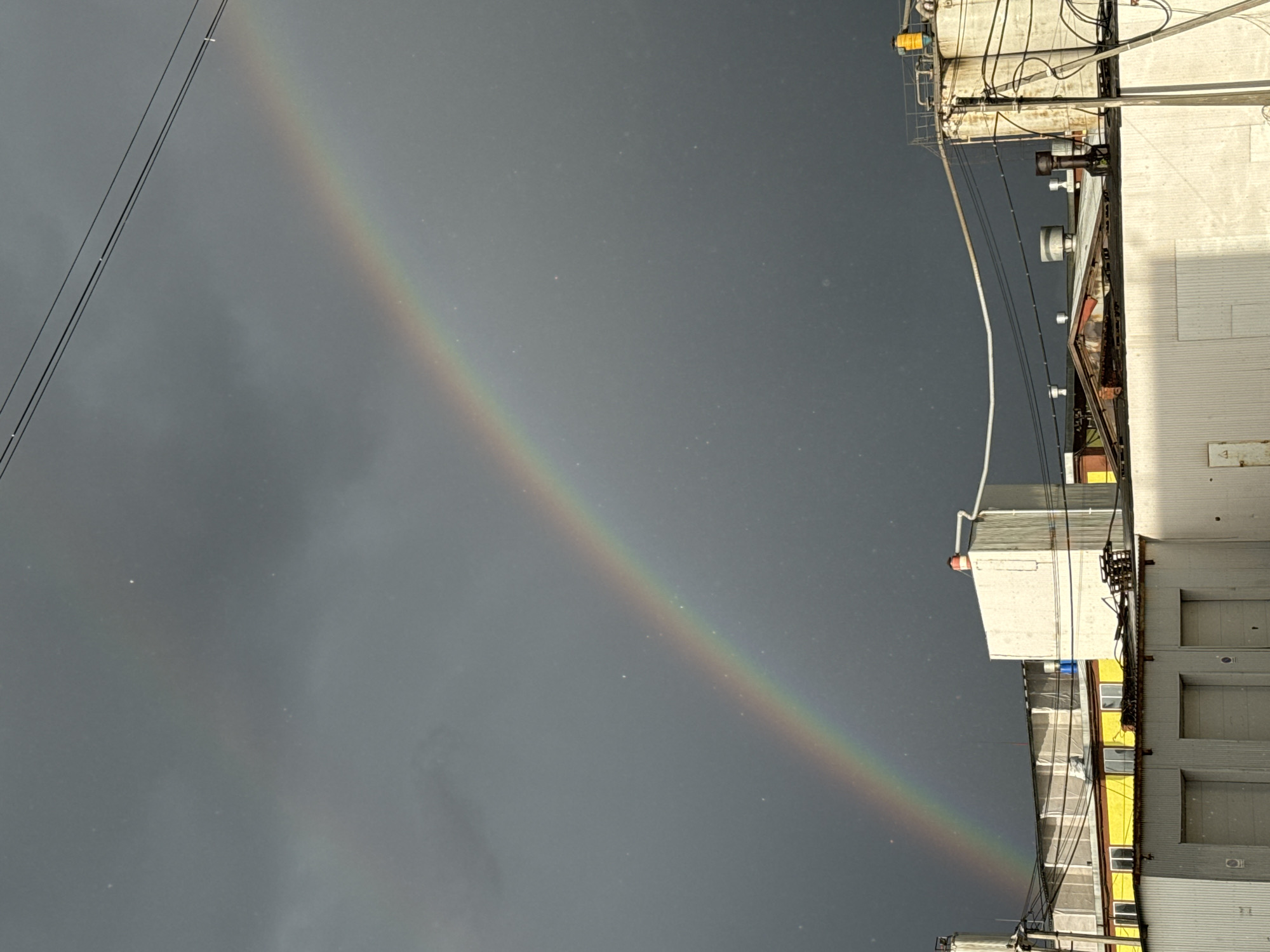 The image shows a rainbow in a grey sky over an industrial area. A building is visible on the right with a corrugated facade and various pipes, wires, and mechanical structures attached to it. The building appears to be in a state of partial disrepair. There are power lines visible in the upper left. The environment appears overcast and possibly rainy, as indicated by the rainbow. The photograph is taken at an odd angle.
There is no information in the image to indicate that this is in the city of Perm, Russia.