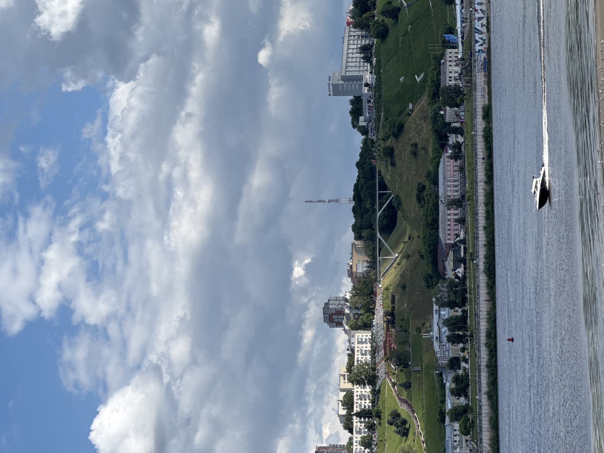 This is a medium-shot photograph of a wide river in the city of Nizhny Novgorod, Russia, under a partly cloudy sky. In the foreground, the blue river fills the bottom third of the frame, with a white and black motorboat speeding across its surface, leaving a foamy wake. The boat is positioned in the lower right quadrant of the image.
The middle ground features a concrete embankment along the river's edge, with some greenery and trees. Above the embankment, a large, steep, green hill rises, dotted with trees and buildings. A white pedestrian bridge spans a section of the hill. Terraced parkland with pathways and seating areas is visible on the hillside. Further up the hill, more buildings are clustered, including some taller apartment blocks and a prominent tall tower with red accents. The Azimut Hotel Nizhny Novgorod, a large, modern grey building with "azimut" signage, is visible on the right side of the hill.
The sky above is a mix of blue sky and large, dramatic grey and white clouds. The lighting suggests it is daytime, possibly mid-afternoon, with some areas of the scene in sunlight and others in shadow cast by the clouds. There are no visible people, though the boat implies human activity. The overall scene depicts a scenic view of the riverfront and the elevated cityscape of Nizhny Novgorod.