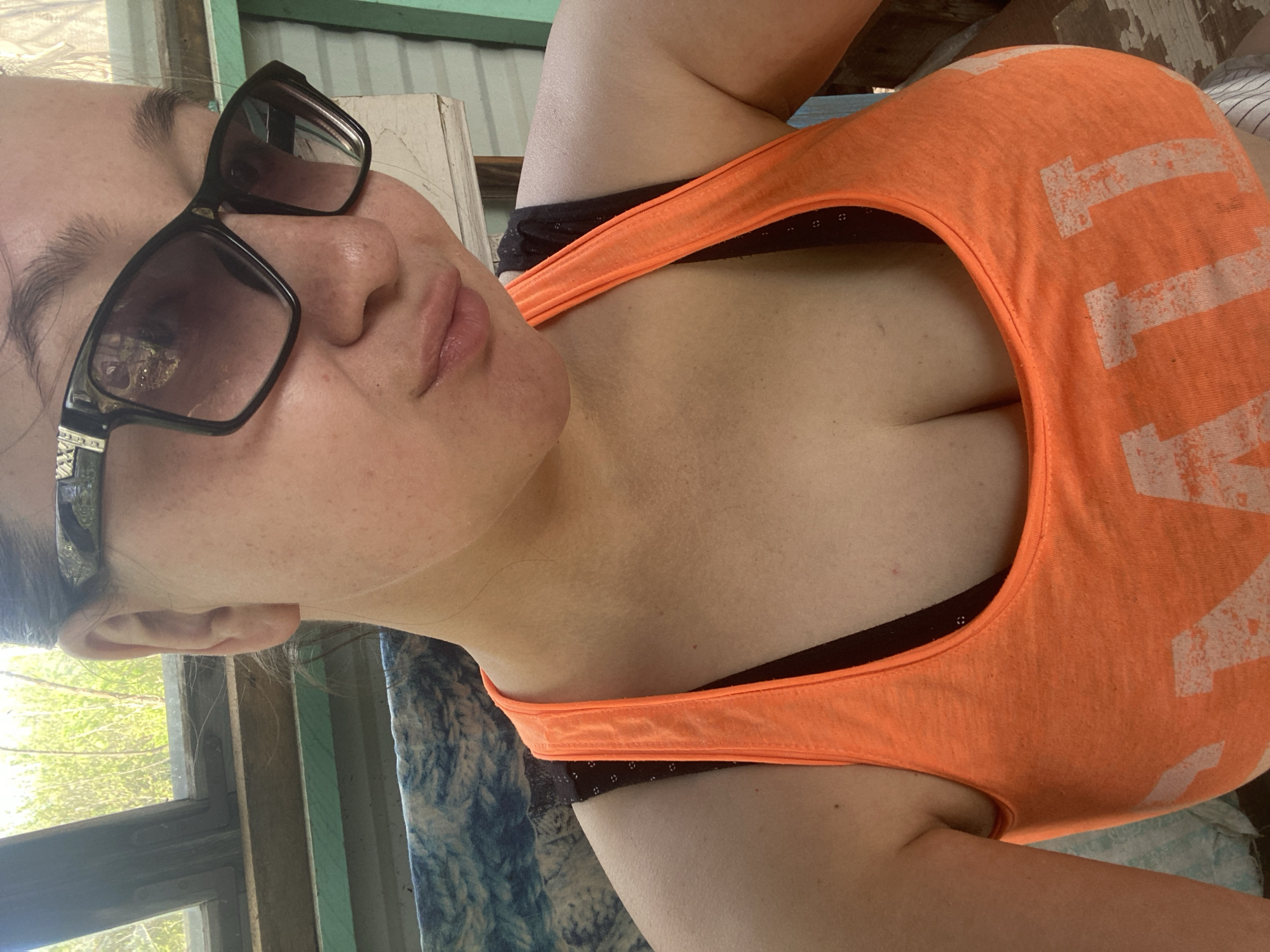 A woman wearing sunglasses and an orange tank top is pictured from a close-up angle. The background includes a window with visible foliage and a textured surface beneath her. The scene is located in the city of Прикамский, Russia.