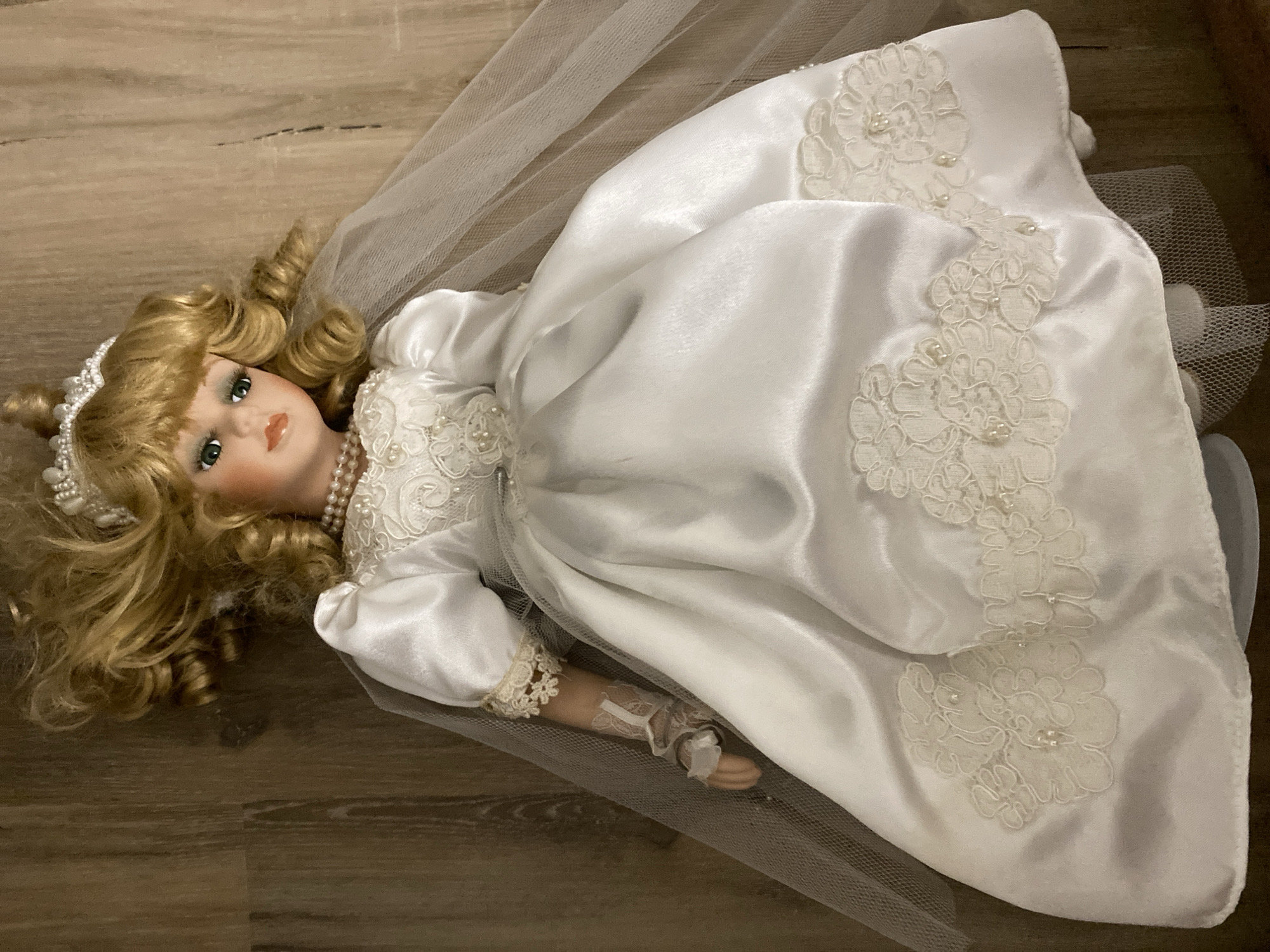 A porcelain doll is lying on a wooden floor. The doll has blonde curly hair, green eyes, and is wearing a white satin wedding dress with lace trim and pearl accents. A sheer veil is draped over the doll's head and shoulders. The doll's right arm is bent at the elbow, and its hand is visible, wearing a lace glove. The doll's dress has a full skirt that extends to the right side of the frame. The wooden floor is visible in the background and foreground, with distinct grain patterns. The lighting is warm and appears to be coming from above, casting soft shadows.