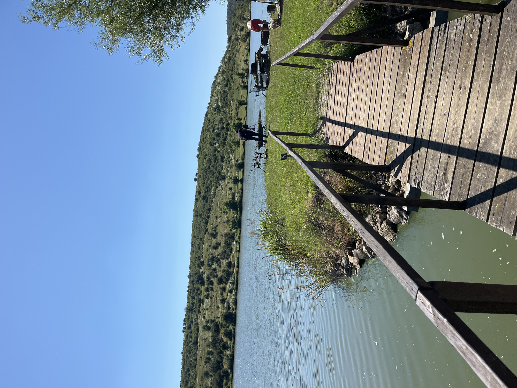 A wooden pier extends over a lake towards a grassy bank where several people are engaged in fishing activities. The lake is calm with a greenish hue, reflecting the clear blue sky. The surrounding landscape is hilly and covered with sparse vegetation and trees. On the bank, fishing rods are set up, and fishing gear is laid out. One person is standing and looking towards the lake, while another is bent over a fishing setup. A third person in red shorts is also visible near the equipment. The pier is made of weathered wooden planks with dark railings. The scene suggests a tranquil day of fishing in an outdoor environment. The location is confirmed to be in the city of Botevo, Bulgaria.