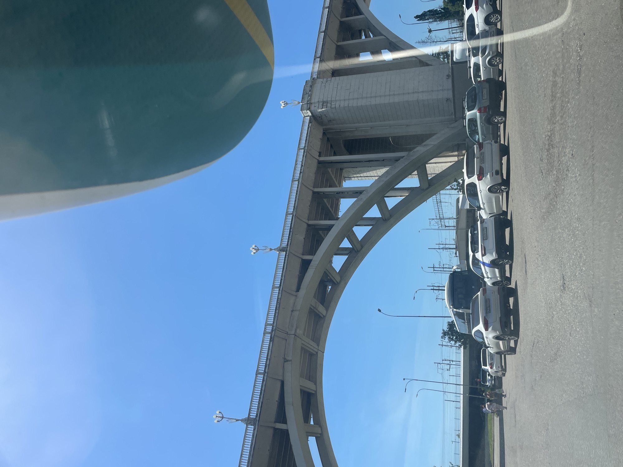 The image captures a large, concrete arched bridge stretching across the frame under a clear, bright blue sky on a sunny day in Sochi, Russia. The bridge, likely a road or railway overpass, exhibits a robust structural design with multiple arches and pillars, and is topped with a railing featuring distinctive multi-bulb lampposts at regular intervals.
Below the bridge and to the right, a line of cars, predominantly white and light-colored, is parked or slowly moving along a paved road. Several visible Russian license plates confirm the location, with the "193 RUS" region code indicating Krasnodar Krai, where Sochi is situated (e.g., "M343CA 193 RUS", "B142KA 193 RUS", "H668PC 193 RUS", "E042CA 193 RUS"). In the distance behind the vehicles, electrical wires and utility poles are visible against the sky. A few indistinct figures of people can be seen walking on a sidewalk or grassy area adjacent to the road.
In the upper left portion of the image, a large, smooth, dark teal or green object with a thin yellow stripe occupies the foreground, suggesting the photo was taken from inside or very near a vehicle or another large structure, potentially a bus or tram. The bright conditions and lack of clouds indicate it is daytime with good weather.