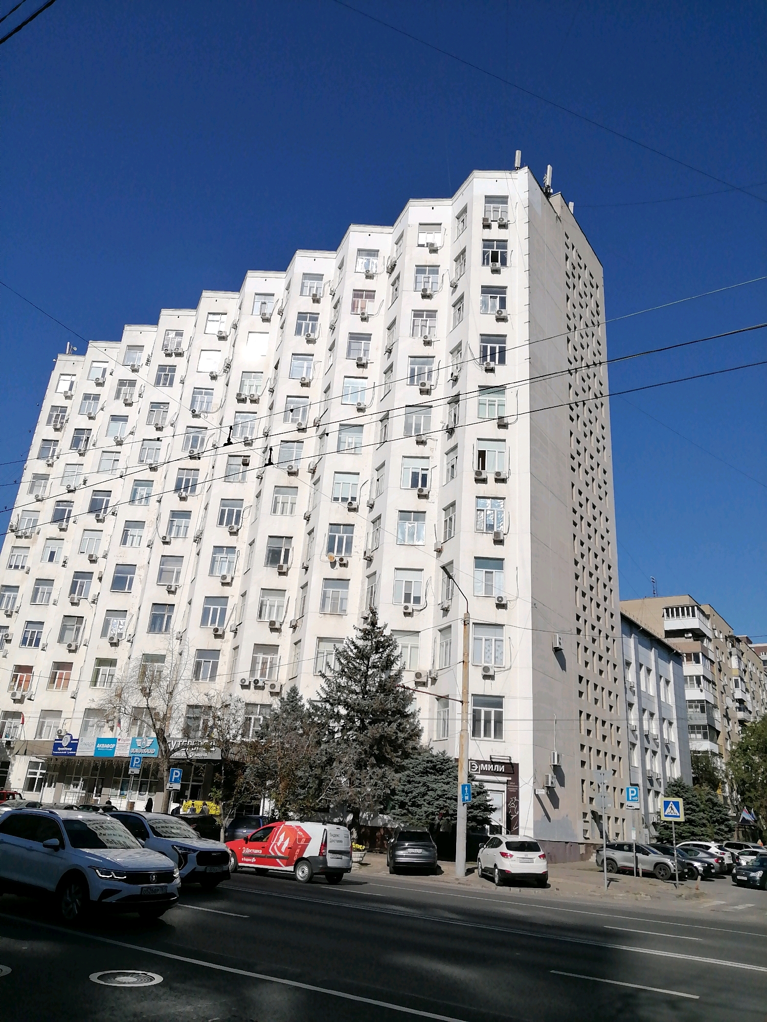 In the city of Rostov-on-Don, Russia, on a clear, sunny day, a large, white, stepped apartment building dominates the frame, its facade covered in numerous windows and air conditioning units. The building is situated on a street with a road in the foreground, where several cars are parked and driving. To the right of the main building, a slightly shorter, beige building with balconies is visible. The sky is a vibrant blue, with power lines crisscrossing the upper portion of the image. Trees, some coniferous and others deciduous, are scattered around the buildings and along the street. The scene captures a typical urban streetscape.