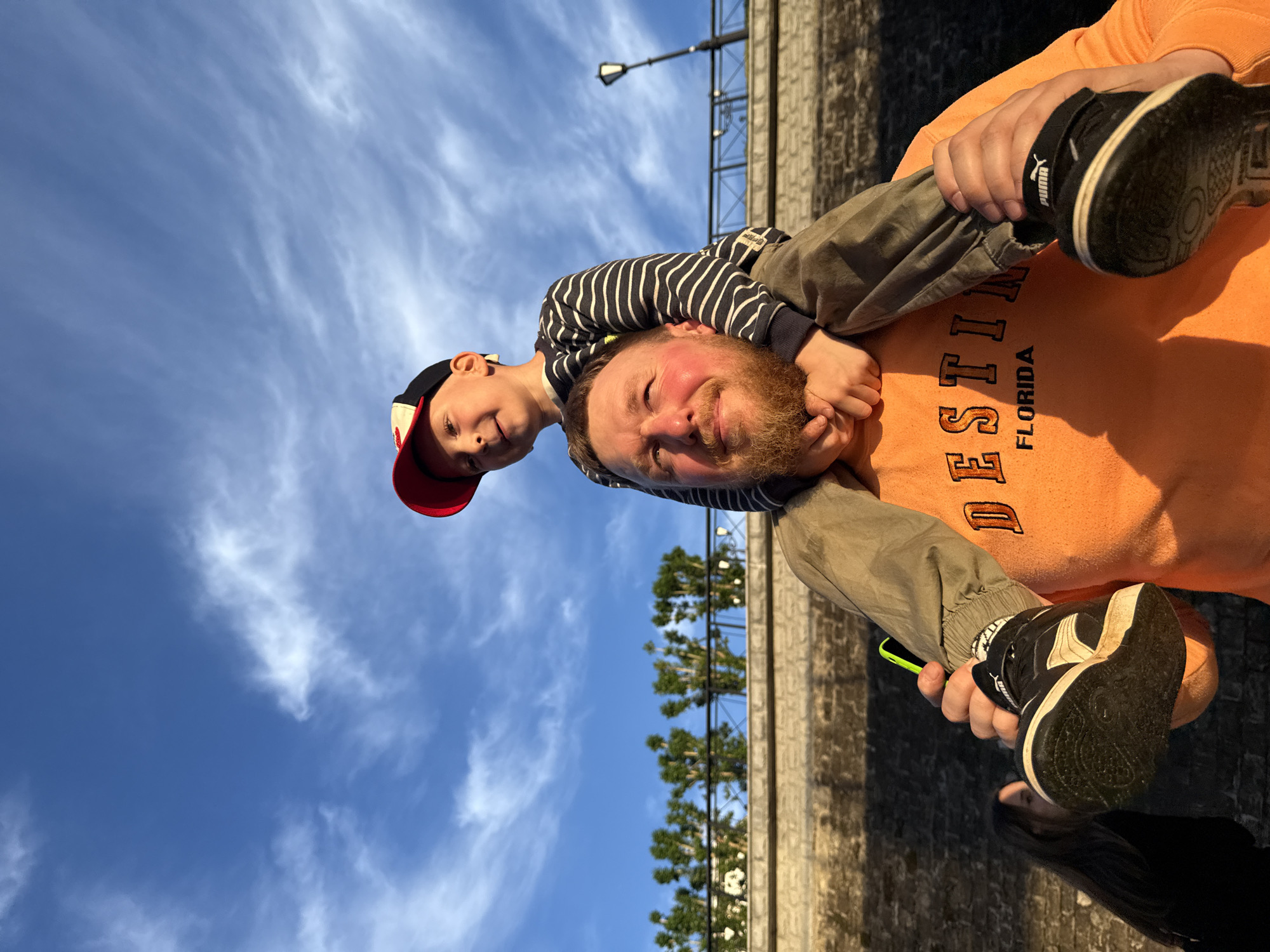 A low-angle medium shot captures a father carrying his young son on his shoulders. The father, with a red beard and wearing an orange sweatshirt, looks upwards with a slight smile. His son, wearing a striped long-sleeved shirt and a red and black baseball cap, looks forward with a grin. The father's hands are around his son's legs, and the son's feet, wearing black Puma sneakers, are visible on his shoulders. In the background, a brick wall and a row of trees are seen against a bright blue sky with wispy clouds. A lamppost is visible on the right side of the frame. A woman with dark hair is partially visible in the lower left corner of the image. The overall setting suggests an outdoor urban environment, possibly a park or embankment in Perm, Russia, during daylight.