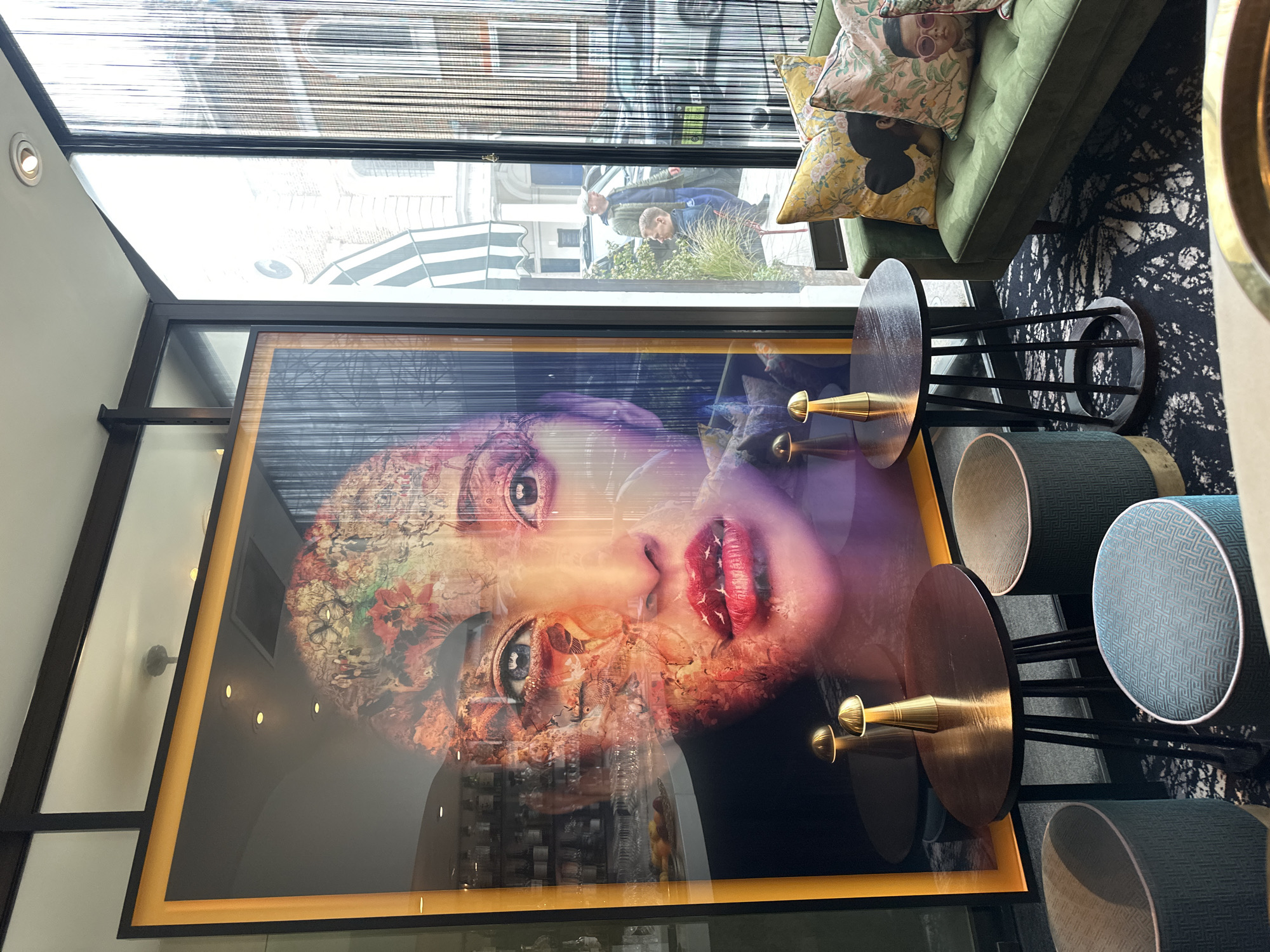 The photograph shows an interior space, likely a cafe or lounge in London, United Kingdom, judging by architectural details reflected in the glass surfaces. The main focal point is a large framed print or painting of a face with surreal elements, including red lips and overlaid images. To the right of the artwork are several small, round tables with stools, featuring a blue, green, and brown color scheme. A green tufted sofa is partially visible in the upper right corner, adorned with decorative pillows.
Through the window or reflective glass, one can discern a street scene with pedestrians and buildings typical of London architecture, like a white building with a striped awning and a gray brick one with small apartments. Black string curtains hang at the top of the window. The room's lighting appears soft and ambient.