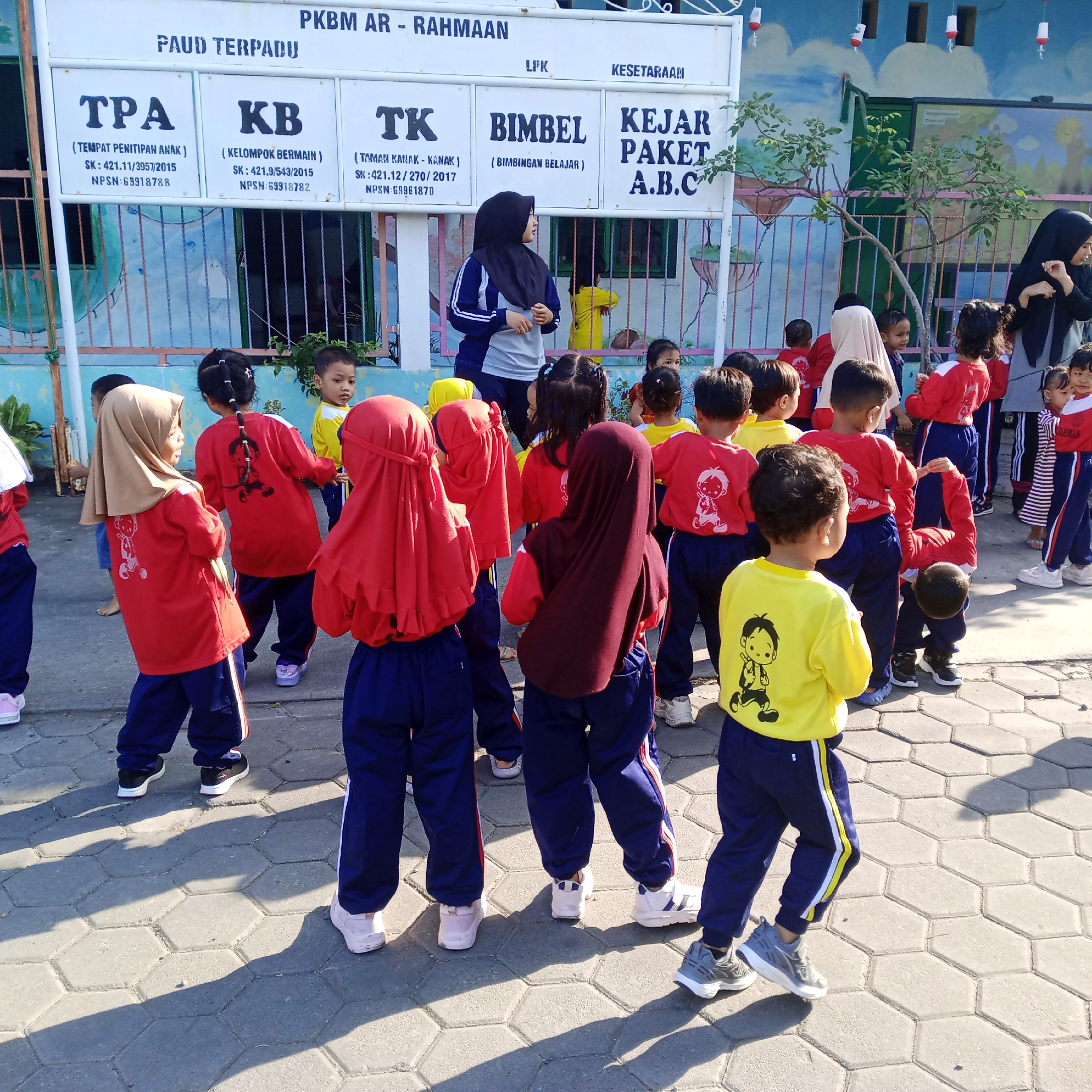 The image depicts an outdoor scene at a school in Rejosari, Indonesia, likely a preschool or kindergarten. A group of young children, dressed in red and yellow shirts and blue pants, are gathered in front of a whiteboard sign. The sign has text in Indonesian, indicating "PAUD TERPADU PKBM AR - RAHMAAN" at the top, with sections for "TPA", "KB", "TK", "BIMBEL", and "KEJAR PAKET A.B.C." Below these headings are various codes and numbers.
Two adult women are present. One, wearing a black hijab and a grey and blue jacket, stands facing the children and appears to be speaking to them or instructing them. Another woman, also wearing a black hijab and a grey top, stands further in the background on the right, observing the scene.
The children are engaged in various activities. Some are standing and looking towards the adult. Others are interacting with each other. One child in a yellow shirt is bending over, as if tying their shoe or reaching for something on the ground. The ground is paved with hexagonal concrete tiles.
The background features a colorful mural on a building wall and a metal fence. The overall environment suggests a schoolyard or an outdoor learning space. The lighting indicates it is daytime, with shadows cast on the ground.