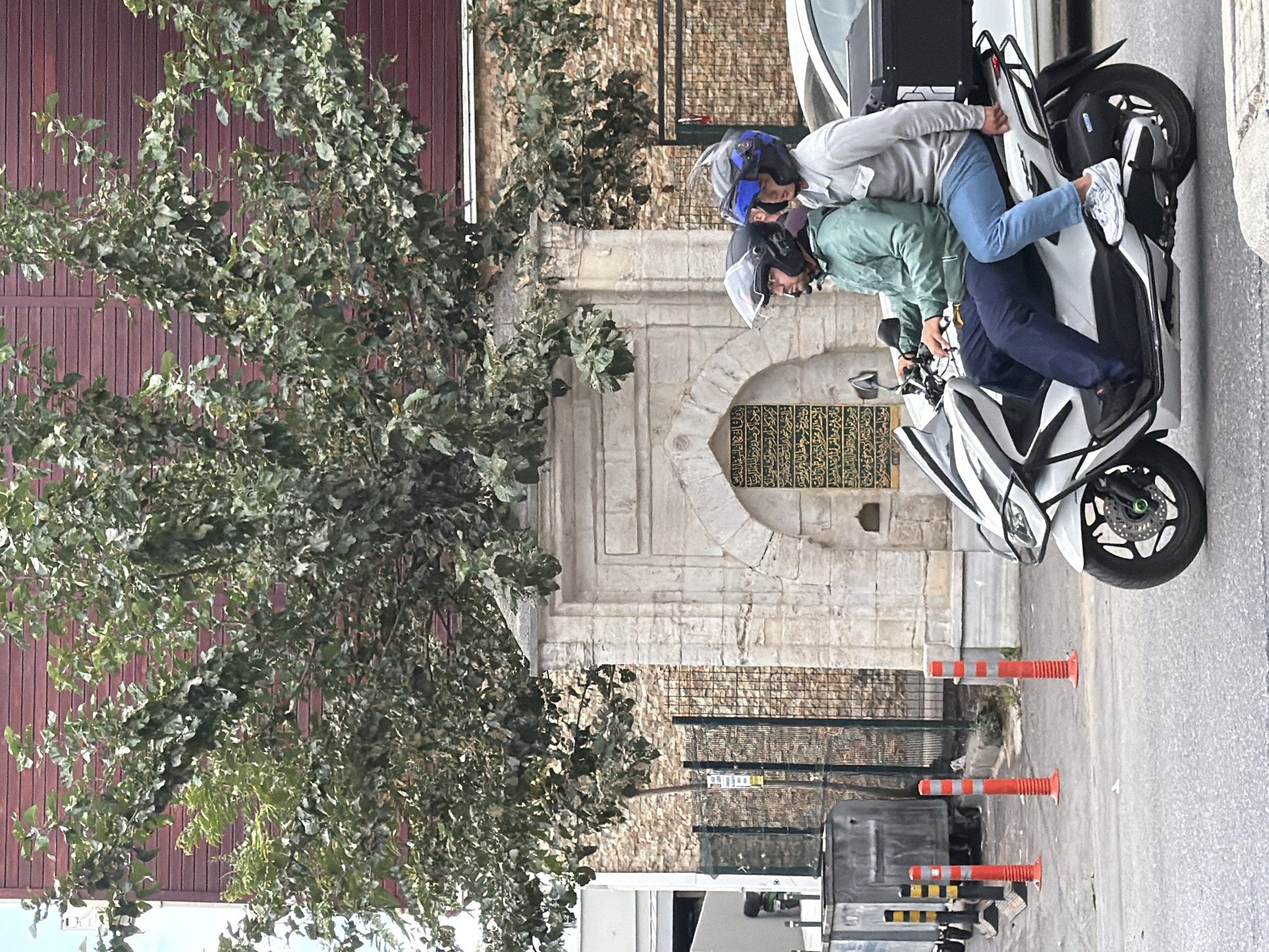 Two individuals, a driver and a passenger, are riding a white scooter in Kadıköy, Turkey. The driver is wearing a green jacket and dark pants, while the passenger is wearing a light gray long-sleeved shirt and blue jeans. Both are wearing helmets, with the driver's helmet being black and the passenger's helmet being blue and black. The scooter has a black storage box mounted on the rear. In the background, there is an ornate stone archway with Arabic calligraphy inscribed on it, likely part of a historical building or monument. A large tree with dense green foliage is positioned above and around the archway. To the left of the archway, there is a metal fence, a brick wall, and a large grey trash receptacle. Several orange traffic cones with reflective stripes are lined up on the side of the road. A portion of a white car is visible to the right of the scooter. The scene takes place on a paved street with a light grey surface. The building behind the tree and archway has a reddish-brown wooden facade.