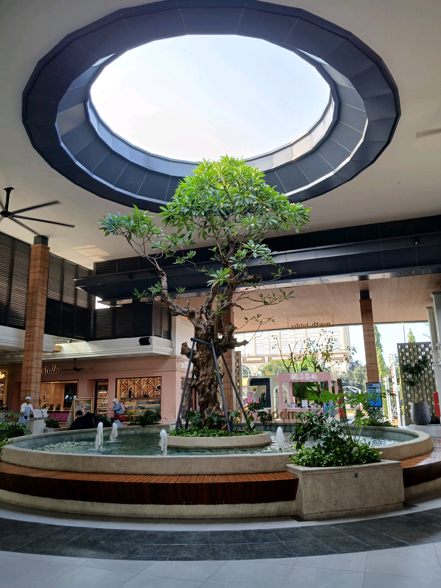 The image shows the interior of a shopping mall atrium in North Jakarta, Indonesia. A large, multi-tiered fountain is the central feature, with a mature tree planted within it. The fountain has several water spouts and is surrounded by wooden benches. Above the fountain, a circular, stepped skylight allows natural light to illuminate the space. In the background, storefronts with signage are visible, along with people browsing and working. Industrial-style ceiling fans are mounted on the ceiling. The overall environment appears modern and designed for a relaxed atmosphere, typical of a high-end mall.