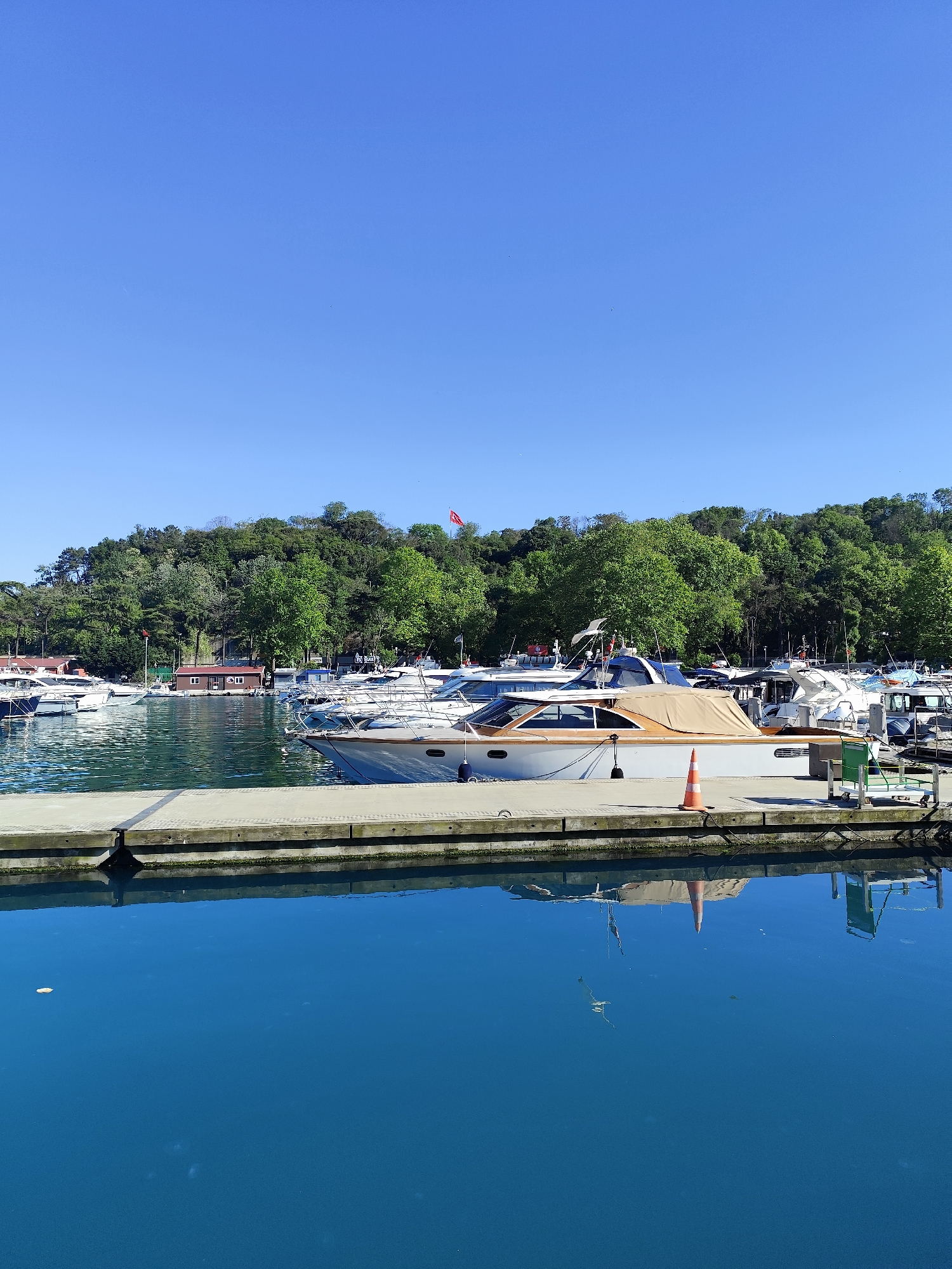 The image shows a marina located in Sarıyer, Turkey, on a clear, sunny day. The scene is dominated by a view of a concrete pier where several yachts are moored. A prominent white yacht with a tan cover sits closest to the viewer, with an orange traffic cone placed on the pier next to it. Other yachts of varying sizes are visible in the background, docked in the marina. The waters of the marina are calm and reflect the boats and pier. Beyond the marina, a green, tree-covered hillside rises, with a Turkish flag visible atop the hill. The sky is a clear, bright blue.