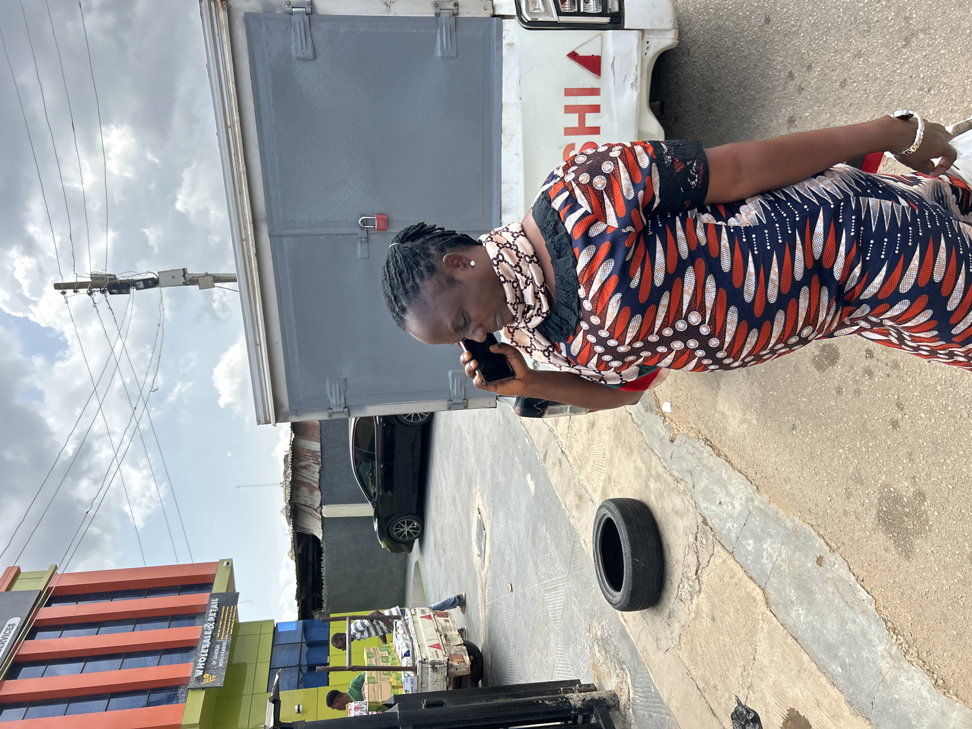 The image captures a bustling daytime street scene in an urban commercial area of Jos, Nigeria, under a cloudy sky.
The central figure is a woman with braided dark hair, engaged in a phone call on a black smartphone held to her ear. She is dressed in a vibrant, patterned gown featuring red, white, blue, and black geometric and circular designs, with black lace accents on the sleeves. A light-colored scarf with a subtle pattern is draped around her neck, and she wears a silver-toned bracelet on her left wrist.
Behind the woman, a white truck with a gray cargo container is parked. The word "ISHI" and a red triangular logo, indicative of "MITSUBISHI," are partially visible on its side. A red padlock secures a latch on the container. On the paved ground beside the woman, a single black tire is resting.
To the left, a multi-story building with distinct orange and green facades and dark windows stands prominently. Legible text on its signage includes "WHOLESALE & RETAIL" and "OF GENERAL MERCHANDISE," pointing to its commercial function. Another partial sign mentions "SERVICES." Further into the background, a black car is parked, and at least two men are visible near what appears to be a vendor's cart or a loaded vehicle, suggesting ongoing trade. Overhead, a utility pole supports multiple power lines and associated electrical equipment against the overcast sky. The ground is paved, exhibiting visible cracks and wear. The overall scene conveys an active, typical day in a commercial part of the city.