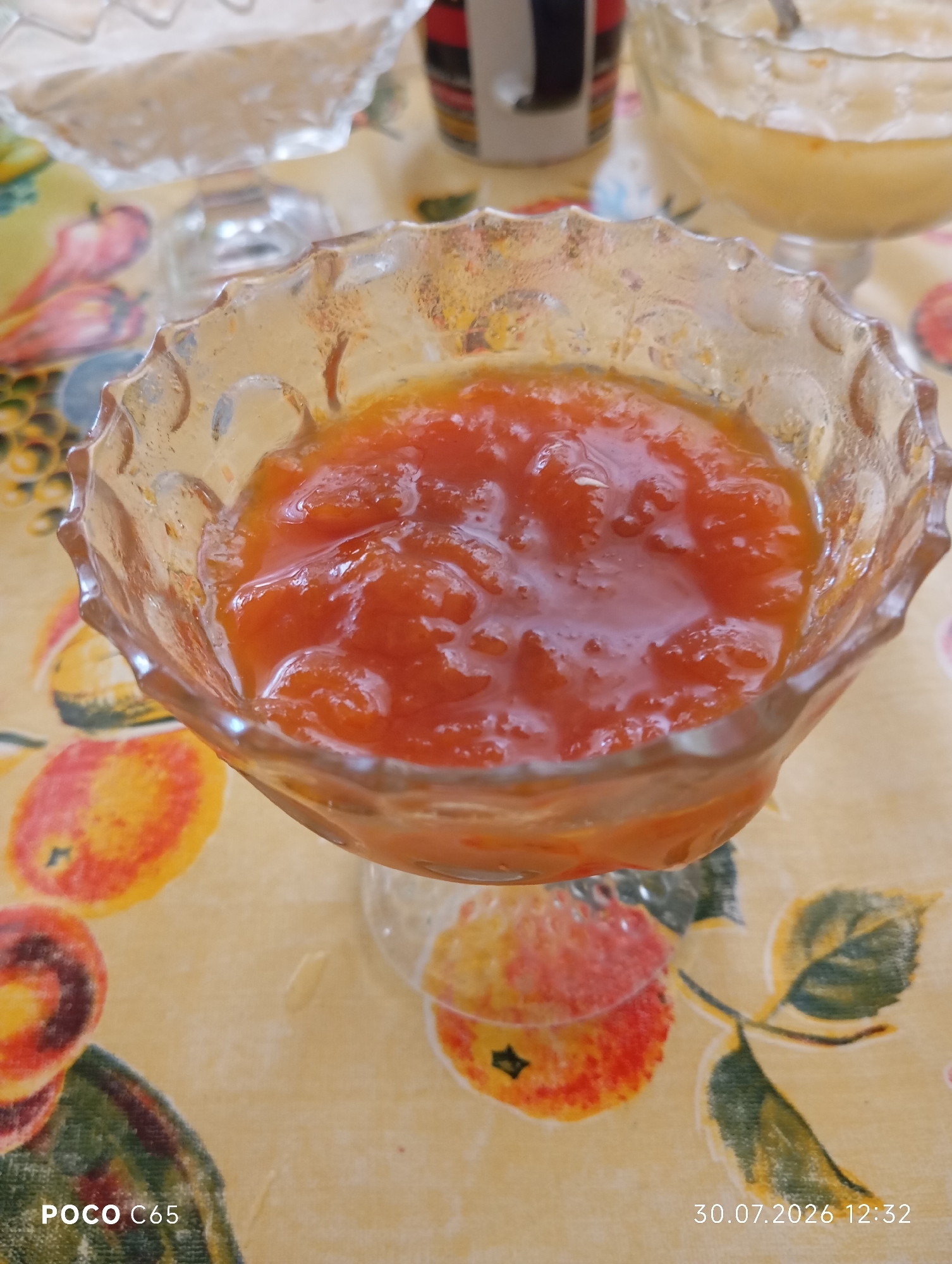 A close-up, eye-level shot shows a glass dessert dish filled with a vibrant orange jam or compote. The dish is ornate, with a scalloped rim and textured glass. The jam is chunky, with visible pieces of fruit. The dish sits on a tablecloth with a yellow background and a pattern of colorful fruits, possibly apples, and green leaves. In the background, out of focus, other dishes containing food items are visible, one appearing to hold a lighter-colored dessert. The lighting is soft and natural. The timestamp "30.07.2026 12:32" is visible in the bottom right corner, and "POCO C65" in the bottom left. No people or specific location within Beruniy, Uzbekistan are identifiable from the image.