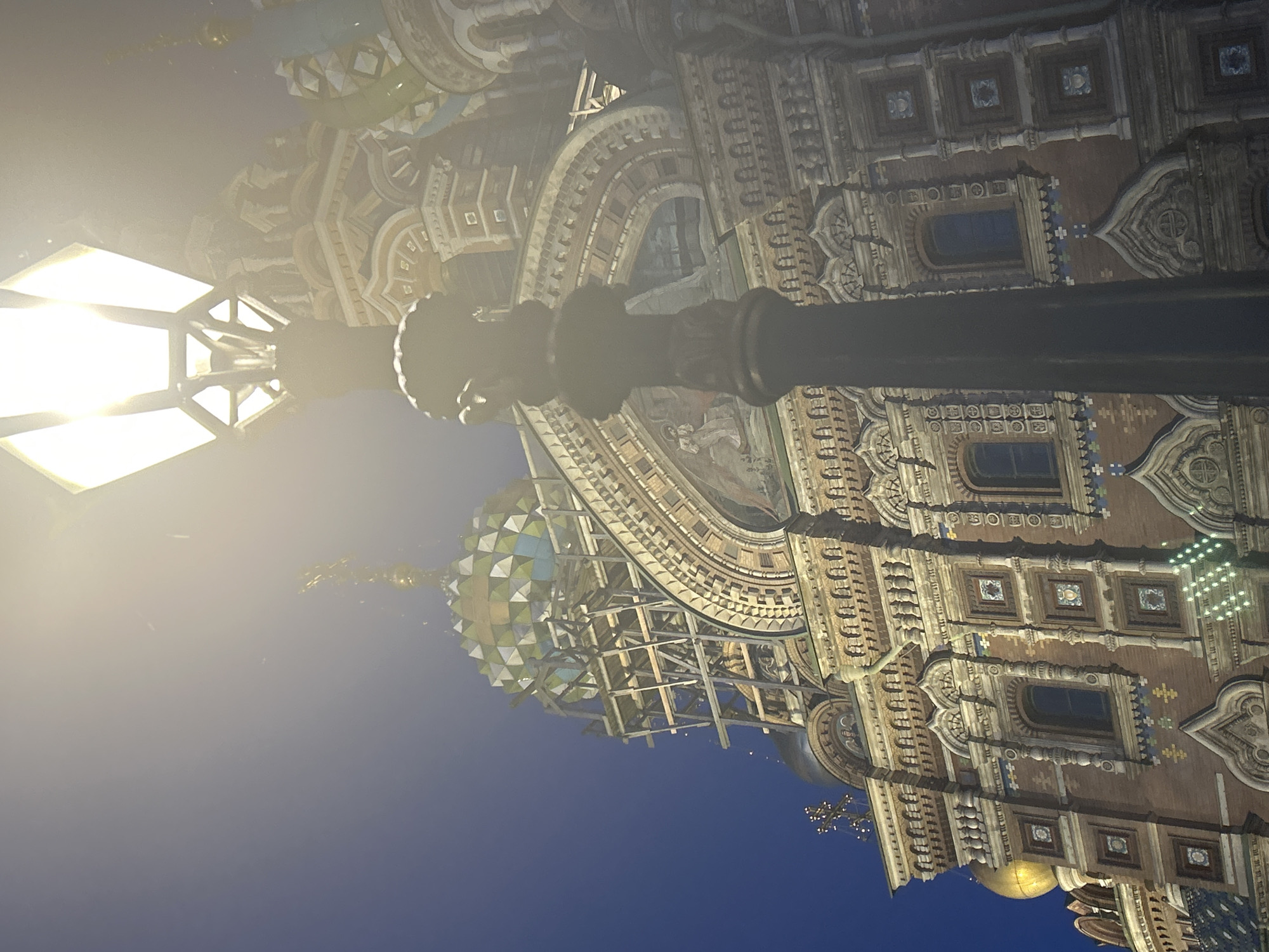 An outdoor, low-angle, eye-level shot captures a street lamp in the foreground against the backdrop of a richly decorated building. The street lamp is a dark, ornate, cast-iron fixture with a hexagonal, luminous lantern at its apex. The building, likely a church or cathedral, displays intricate architectural details, including mosaic domes, patterned brickwork, arched windows, and decorative friezes. One dome appears to be partially under construction, with scaffolding visible. The sky is a deep blue, and a bright light emanates from the street lamp, creating a lens flare effect that washes out parts of the image. The lighting suggests it is either dawn or dusk, or the scene is artificially lit. There are no people visible in the image.