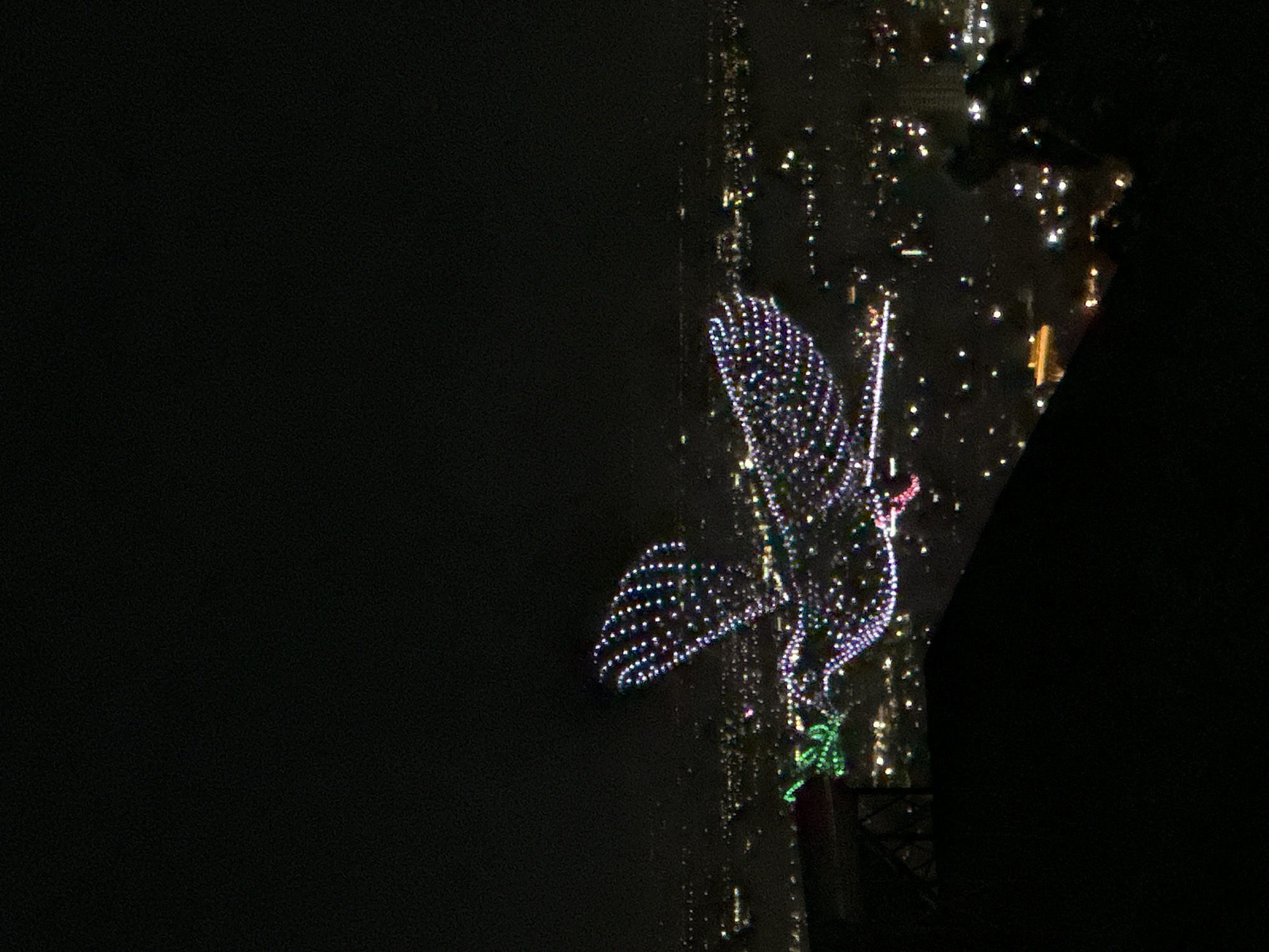 The image captures a nighttime aerial view of a city, dominated by a large, illuminated dove figure in the mid-ground. The dove, composed of numerous small, multi-colored lights, primarily white, purple, and pink, appears to be in flight with its wings spread. A green illuminated branch is visible within its beak area.
Below and extending into the background, the city of Yerevan, Armenia, is visible as a dense network of scattered white, yellow, and red artificial lights, indicating urban development and infrastructure. The horizon is indistinguishable due to the darkness and city lights. The overall atmosphere is dark, with ambient light emanating solely from the illuminated dove and the city below.
In the immediate foreground, at the bottom left, a dark, indistinct structure with a visible red surface (possibly a table or platform) partially obstructs the view. The bottom right shows dark, amorphous shapes, likely part of the terrain or a building from which the photograph was taken. The perspective suggests the image was captured from an elevated position overlooking the city, possibly from a viewpoint such as the Cascade complex. No individuals are discernible in the scene.