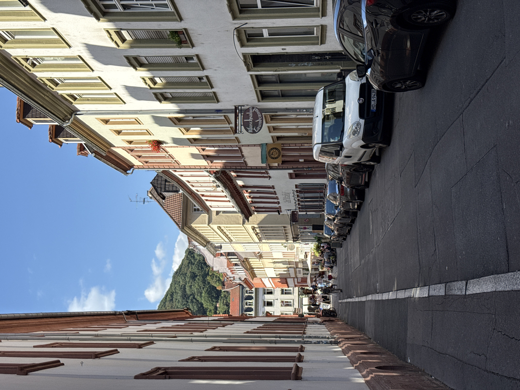 The image shows a narrow street in Heidelberg, Germany, lined with multi-story buildings with colorful facades. The street is paved and there are parked cars on the right side of the road. Several people are walking and sitting at outdoor tables in front of some of the establishments on the left side of the street. The buildings have a traditional European architectural style, with windows and balconies. A lush green hill can be seen in the background. The sky is blue with a few scattered clouds. The lighting suggests it is daytime.