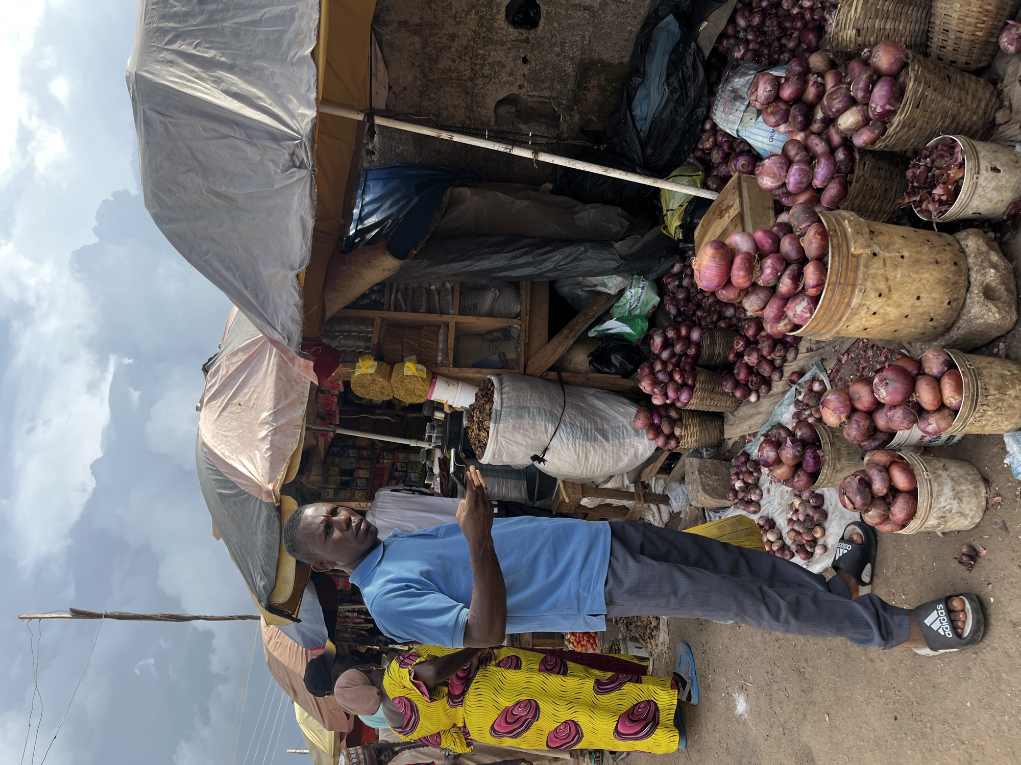 A man in a blue polo shirt and gray pants stands in a market in Rafin Par, Nigeria, holding a mobile phone. He is looking towards the viewer with a neutral expression. Behind him, a woman in a vibrant yellow dress with pink circular patterns is also visible. The market stalls are covered by awnings, and there are piles of red onions displayed in baskets and buckets. The ground is dirt. The sky is cloudy.