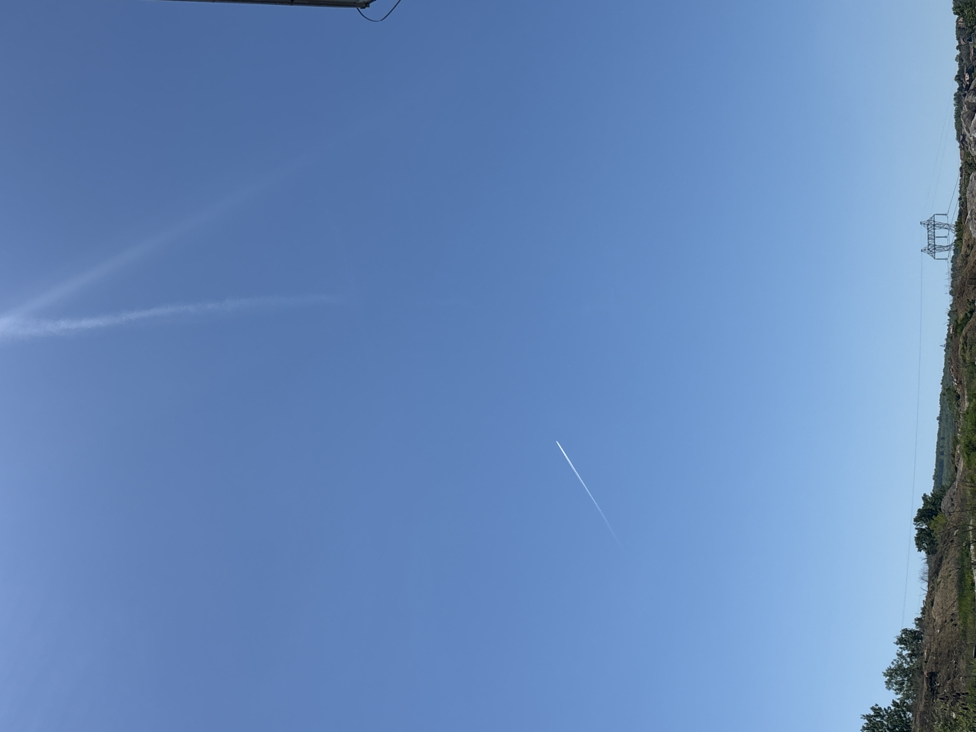 A clear blue sky occupies the majority of the frame. Two visible contrails indicate the passage of aircraft. The ground is visible on the right side of the frame, showing vegetation and an electrical transmission tower, suggesting an open, possibly rural location. This scene is likely located near Grocka Urban Municipality, Serbia, based on the presence of the electrical tower and the natural landscape. The image shows no people.