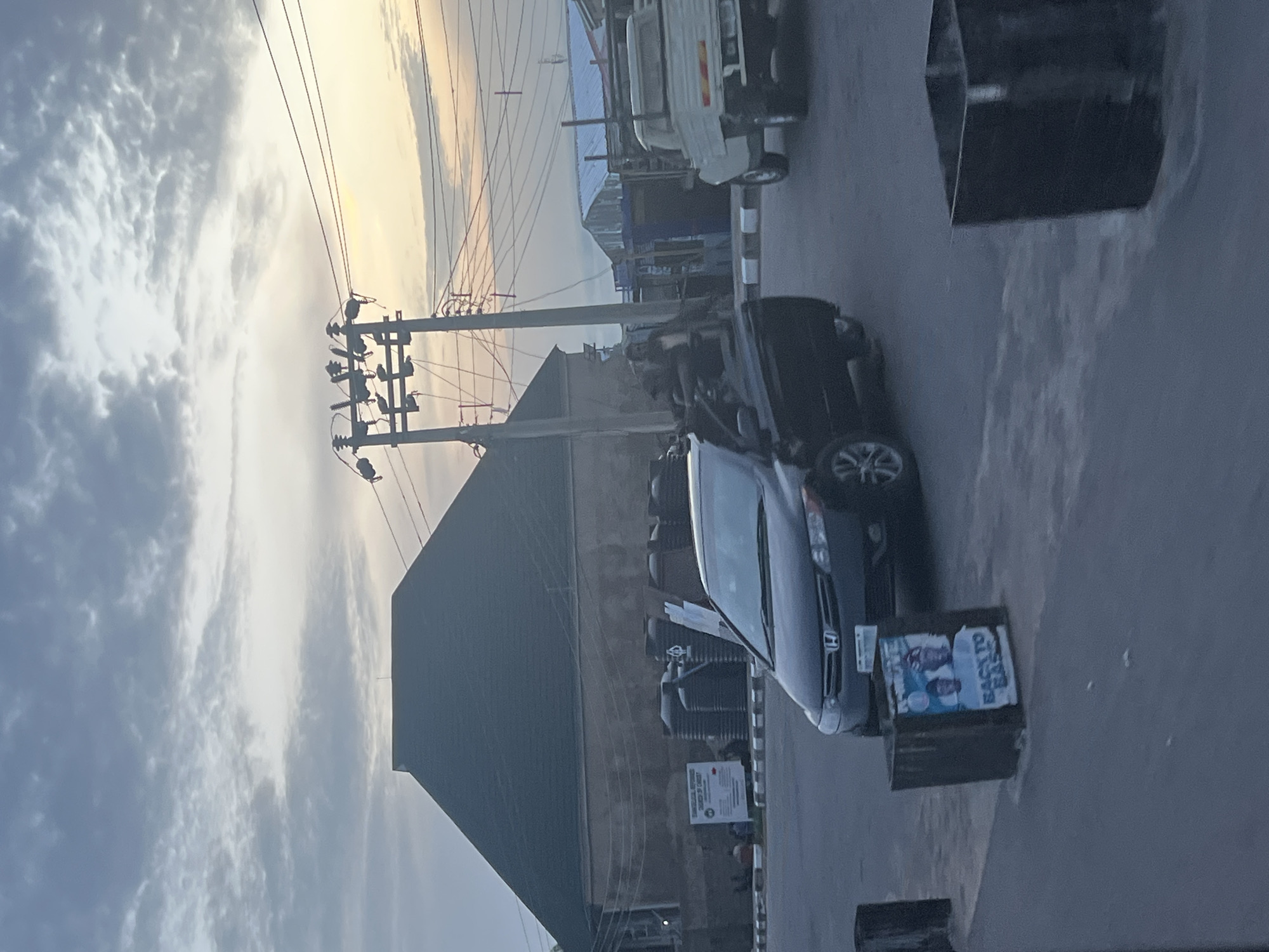 The image captures an outdoor scene, likely a street in Jalingo, Nigeria, during a cloudy dawn. In the foreground, a black sedan is parked, its front passenger door ajar. A poster on a bin in front of the car displays two individuals with the text "BACK TO BASICS". Behind the sedan, a white truck is parked. A building with a dark, pitched roof dominates the mid-ground, with several large black water tanks visible to its right. Power lines and utility poles are prominent in the sky, with the soft light of sunrise visible on the horizon. The overall atmosphere is subdued, with a focus on urban infrastructure and everyday life.
