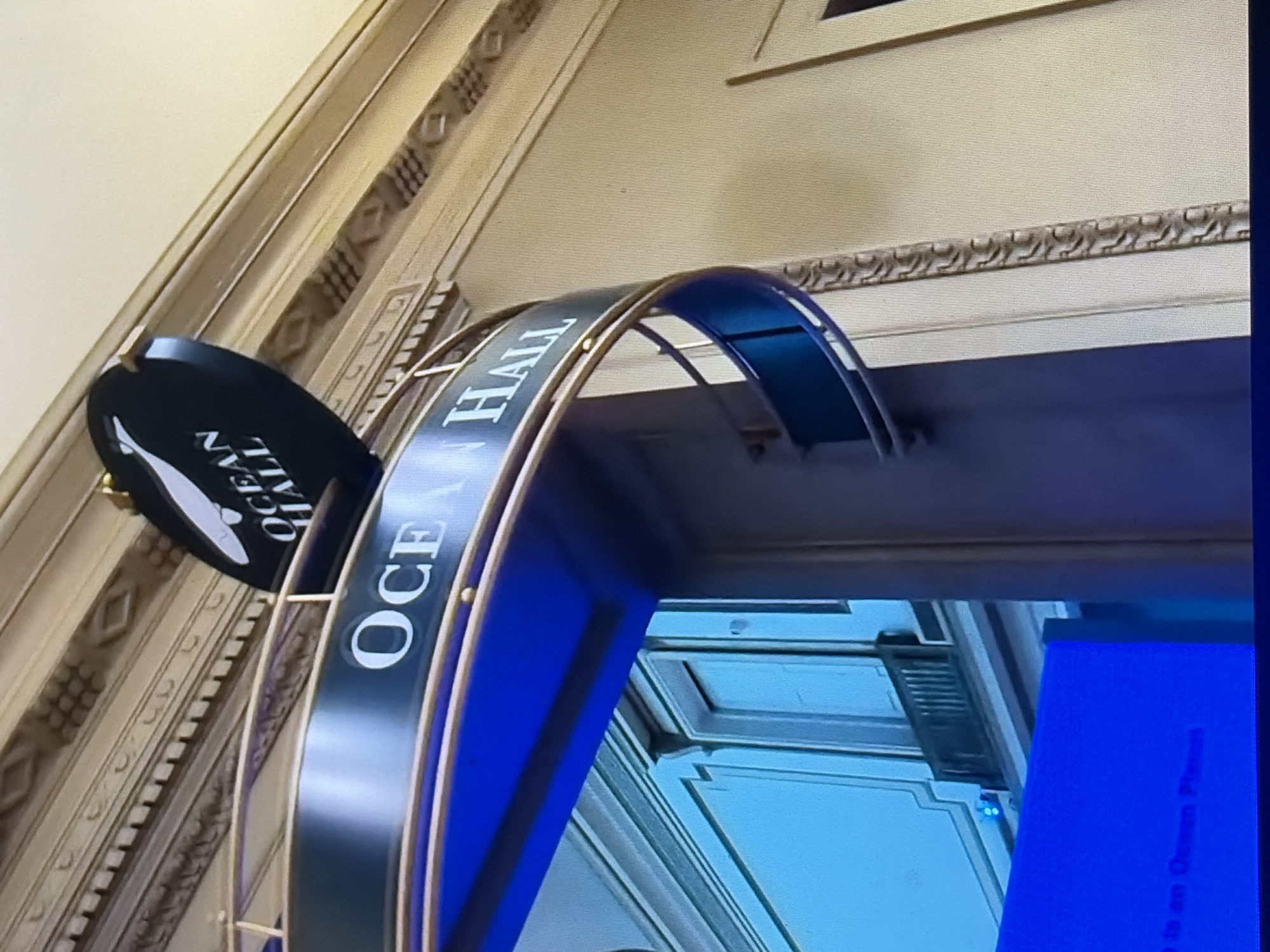 The image shows a sign for "OCEAN HALL" above an entranceway. The sign is curved and appears to be made of dark material with white lettering. There is a whale graphic on the side of the sign. The entranceway has a blue structure beneath it, and a decorative railing. The background shows ornate architectural details on a wall and ceiling. The setting appears to be indoors, possibly a museum or a public building. The overall impression is of a grand or formal entrance.