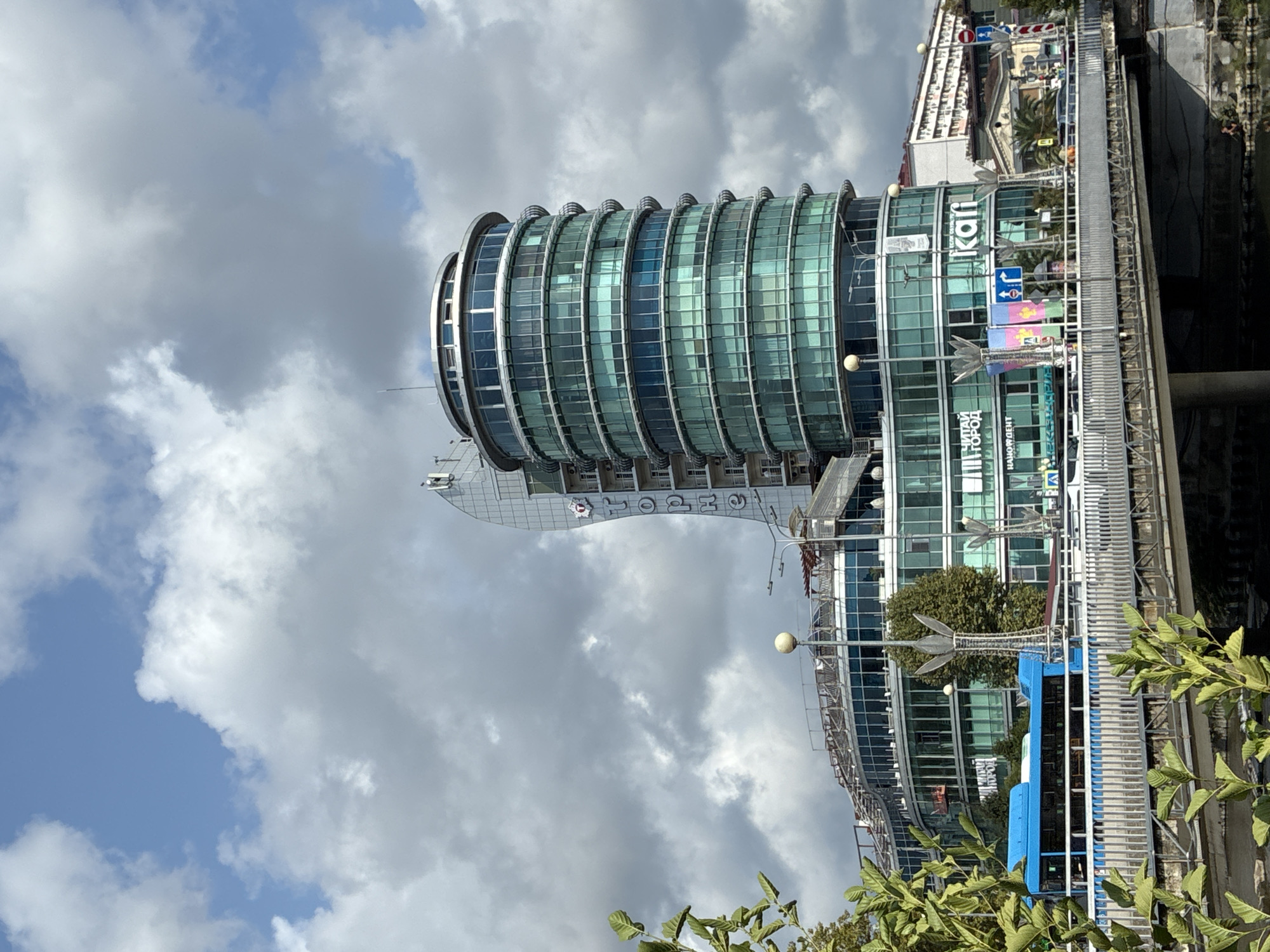This is a medium-shot, eye-level, outdoor, daylight, vertical, photographic image of a modern building complex in Sochi, Russia. The main building is cylindrical with a glass facade and numerous floors. To the left and slightly behind it is another building with a curved glass facade, and a tall, slender, white tower with vertical text. The text on the tower, in Cyrillic, appears to read "TOPHЕ". Signs on the lower floors of the main building are also in Cyrillic and include the word "KAFI". A bright blue bus is visible on a bridge in the foreground, crossing over a body of water. The bridge is made of metal and concrete. Lush green foliage from trees frames the left side of the image. The sky is a mix of blue and white clouds. The overall impression is a contemporary urban scene.