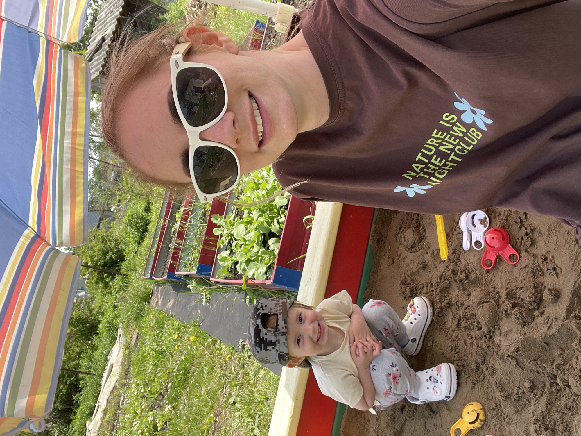 The image shows a woman and a young child in a sandbox. The woman is wearing a brown t-shirt with the text "NATURE IS THE NEW NIGHTCLUB" and white sunglasses. The child is wearing a beige t-shirt, floral leggings, and a camouflage hat. The sandbox contains various plastic toys. An umbrella stands to the left, providing shade. There is a garden patch next to the sandbox, with green plants growing. The location is identified as Yasnoe, Russia.