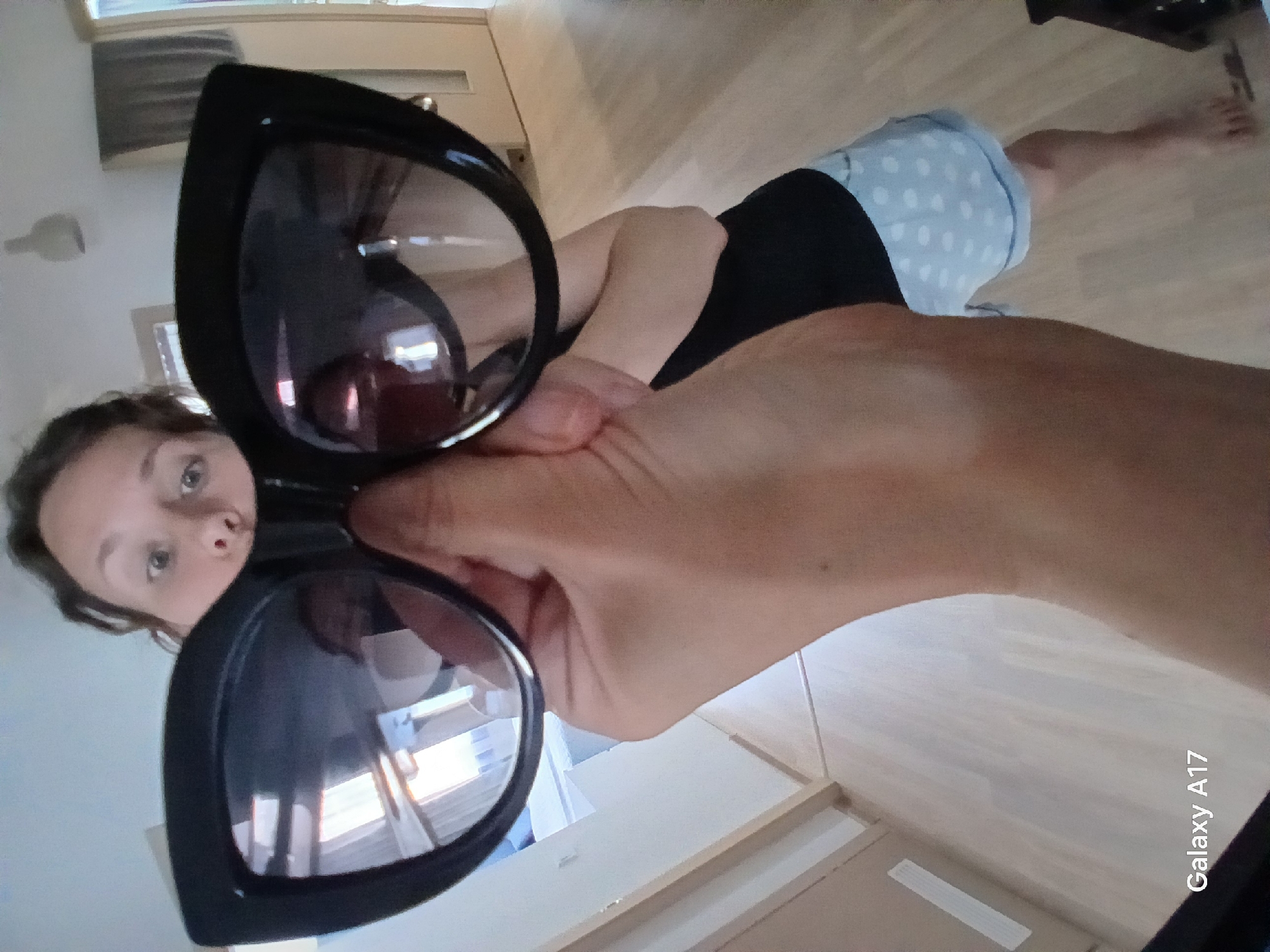 A woman is holding up a pair of black sunglasses in front of the camera. She has light skin, and is wearing a black tank top and blue and white polka dot shorts. Her bare feet are visible in the background on a wooden floor. The background includes a window, white walls, and a wooden floor. The bottom right corner of the image displays the text "Galaxy A17". The location is difficult to assess and is not likely to be Yekaterinburg Russia due to architecture and setting.