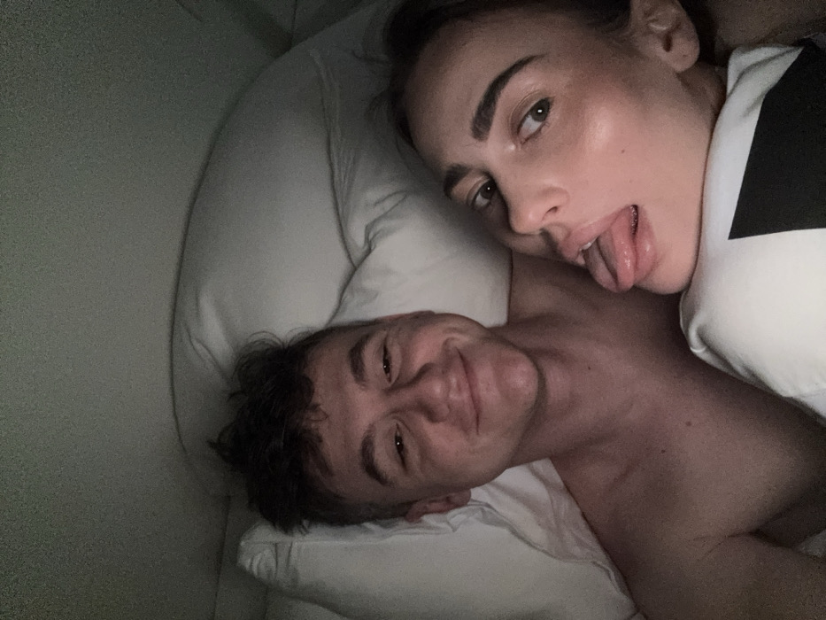 This image depicts two individuals in a dimly lit indoor setting, consistent with a bedroom, located in Venice, Italy. On the left, a young adult male lies on his back, his head resting on a white pillow. He has short, dark, tousled hair and a light complexion, smiling directly towards the camera. His upper torso is visible and appears bare. To his right, a young adult female is positioned alongside him. She has dark hair, likely pulled back, and a light complexion, looking towards the camera with her tongue extended in a playful expression. She wears a white garment featuring a prominent black geometric pattern across the chest. The immediate environment is characterized by white pillows and bed coverings, with the background being indistinct and dark, suggesting low ambient light.
