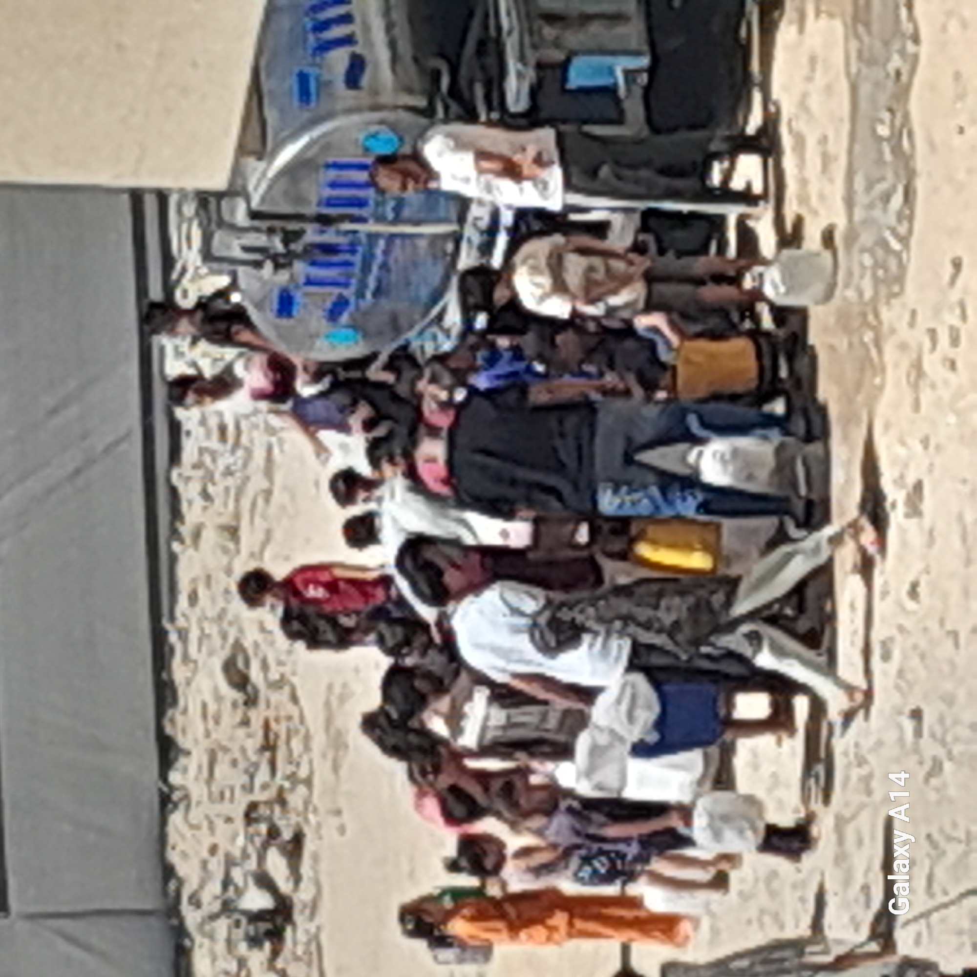 In Khan Yunis, Palestinian Territories, a group of people, including children and adults, are gathered around a water truck. Some individuals are carrying empty containers, likely to collect water. The ground is dusty and uneven, with scattered debris. The overall scene suggests a distribution of essential resources in a community setting. The water truck, a large metallic cylindrical vehicle, is a prominent object in the image, indicating its importance as a source of water. People are dressed in casual clothing, and their expressions are not clearly discernible due to the image quality. The background appears to be an open area with some structures or tents, further suggesting a community or temporary settlement.