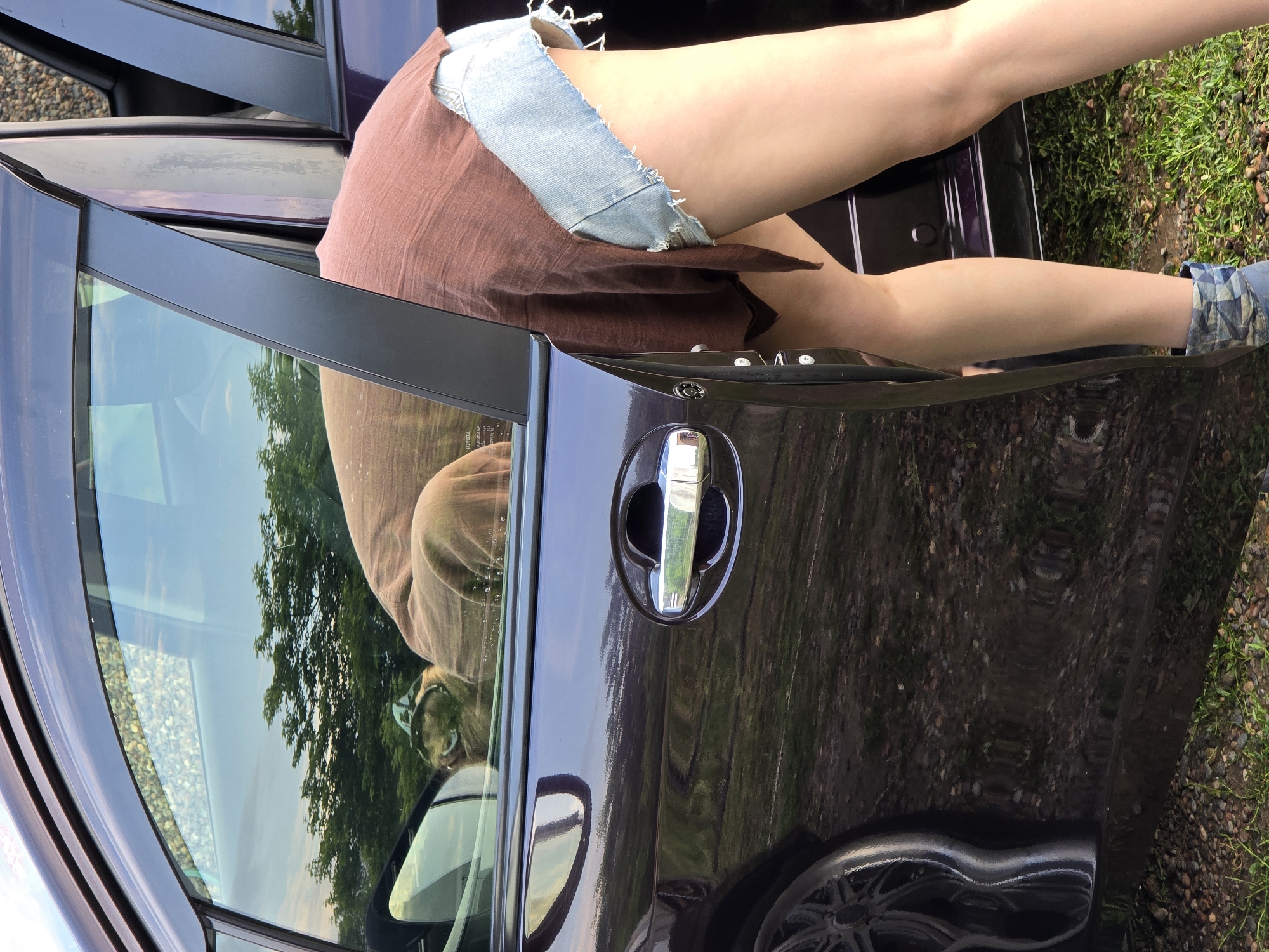 A person is partially visible, leaning into the open driver's side door of a dark-colored vehicle. They are wearing denim shorts and a loose-fitting brown top. The interior of the car is visible through the window, with a reflection of trees and the sky. The exterior of the car shows reflections of the surrounding environment, which appears to be a grassy area with gravel. The scene is likely outdoors, possibly in a rural or suburban setting. There is no information to suggest the location is in Ермолаево, Russia.