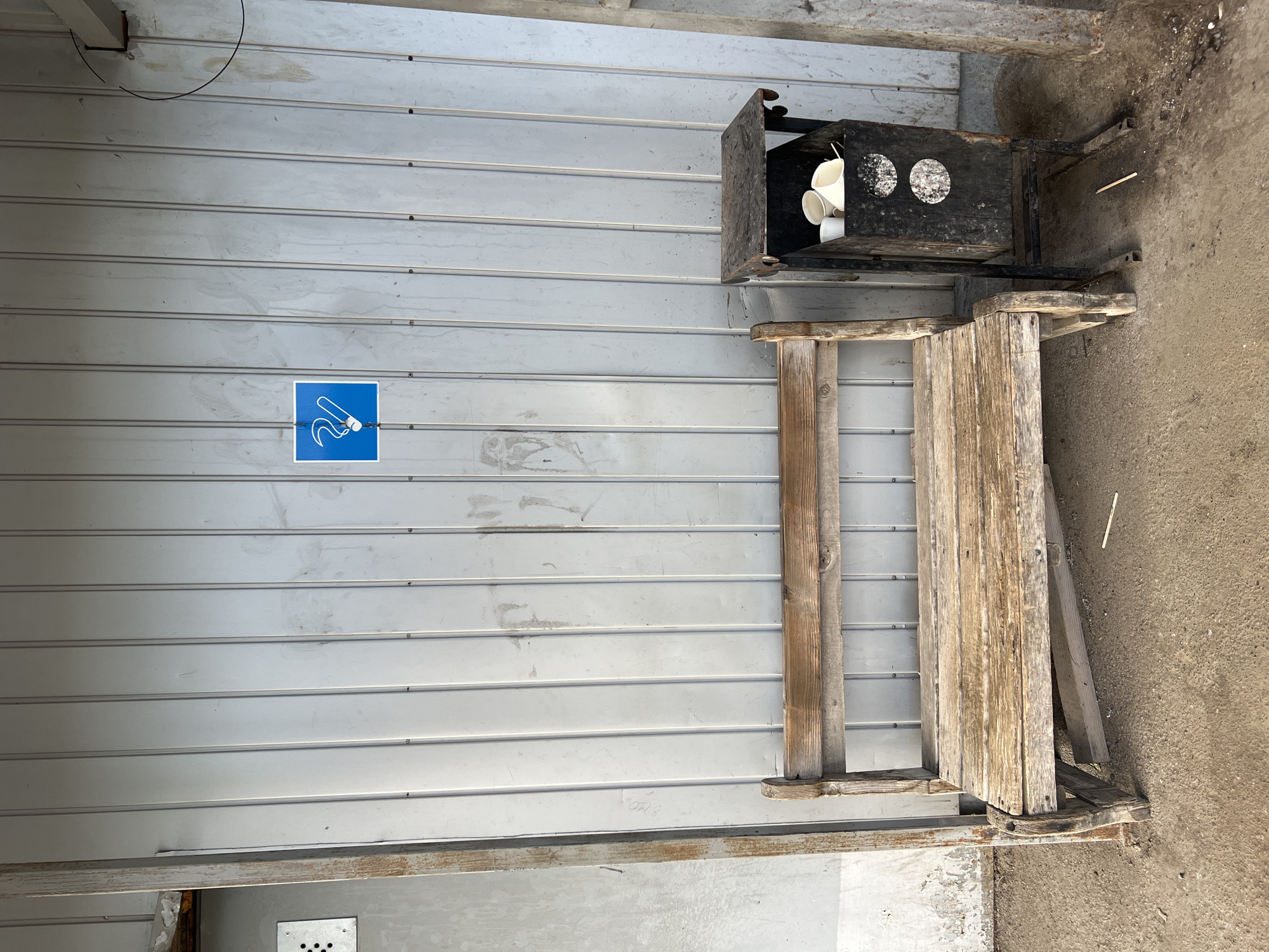 The image depicts an outdoor, static scene featuring a designated smoking area against a weathered, light gray corrugated metal wall. The image lacks specific visual identifiers or textual information to confirm its location as Izhevsk, Russia.
A blue square sign with a white graphic of a lit cigarette and curling smoke is affixed to the wall, positioned in the upper half. The metal wall exhibits vertical panel lines, visible fasteners, and various streaks, smudges, and areas of grime. A faint numeral sequence, possibly "8110," is visible in graphite or light pencil on the left side of the wall. On the far left, a vertical metal beam separates the corrugated wall from an adjacent, lighter-colored, textured wall section with a small, square metal grate. A dark, thin cable or pipe is partially visible in the upper right quadrant.
In the foreground, a rustic, weathered wooden bench with a slatted seat and backrest is positioned to the left. Its timber appears aged and worn. To the right of the bench stands a dark, possibly black or dark brown, metal refuse container, which also functions as an ashtray. This container has a small, flat top and a main bin filled with several white, disposable cups and what appears to be cigarette ash and butts. Two faded white circular markings are visible on the front of the bin. The ground beneath the bench and container is a dirty, uneven surface of compacted earth or concrete, with a few small pieces of debris, including a light-colored stick near the base of the ashtray. No individuals are present in the image, and no specific actions are depicted. The overall scene suggests a functional, utilitarian space.