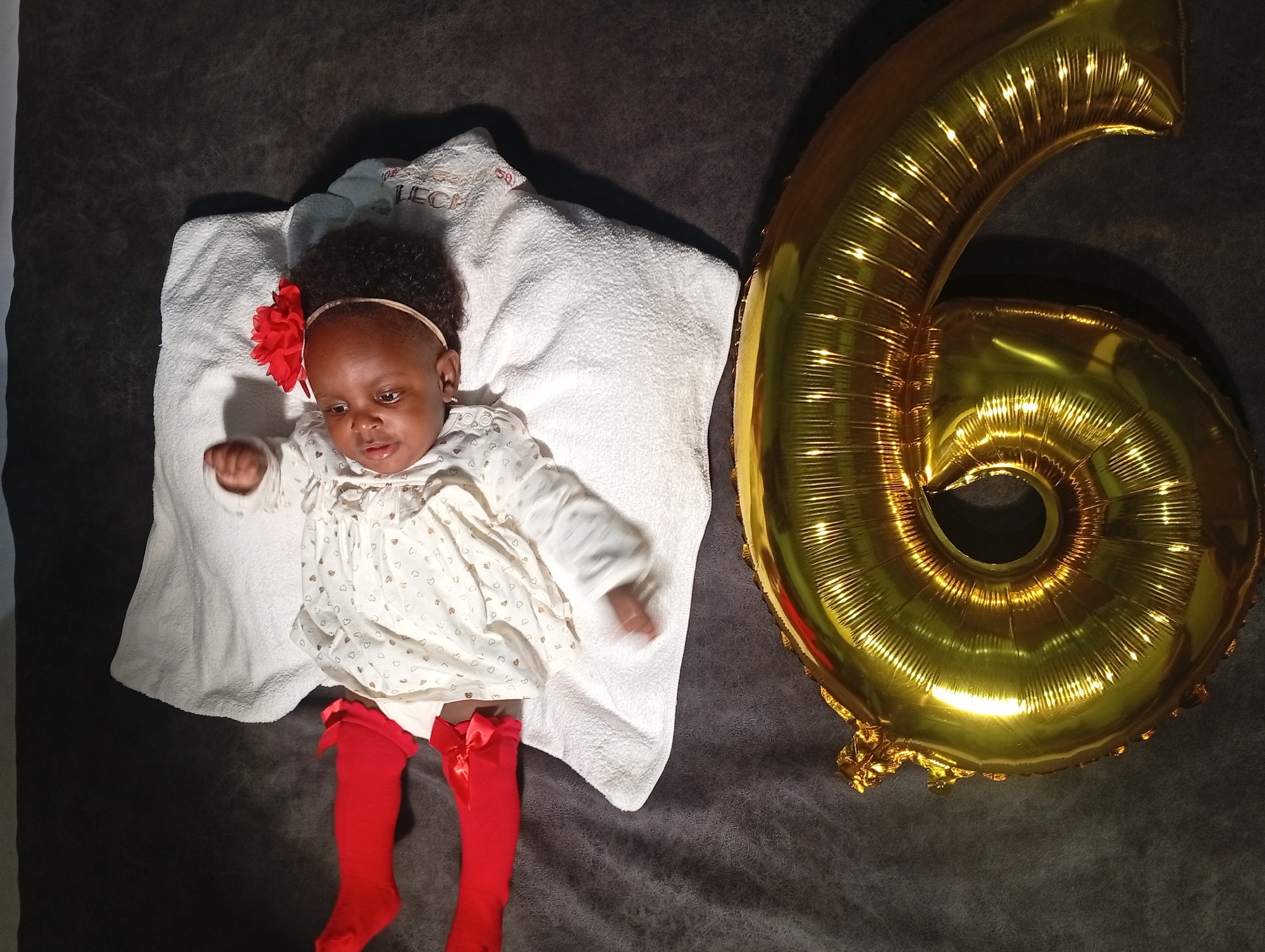 A baby is lying on their back on a white towel, which is itself placed on a dark gray surface, presumably a bed or blanket. The baby is wearing a light-colored dress with small heart patterns, red tights with bows at the knees, and a headband with a red flower. To the baby's right is a large, gold-colored balloon shaped like the number '6'. There's insufficient evidence to conclude the location of the image is Barakin Akawo, Nigeria.