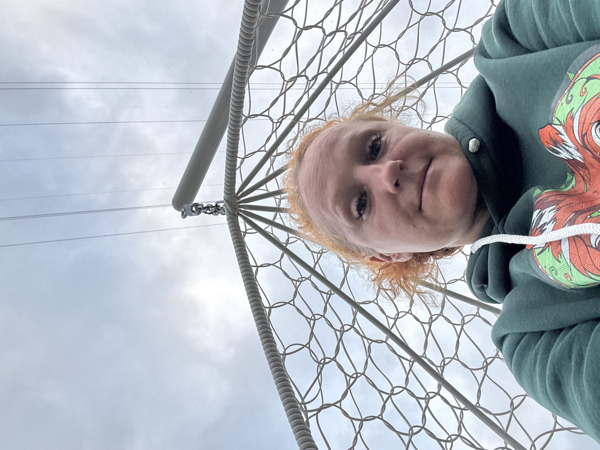 A woman with red hair and a green hooded sweatshirt is photographed from a low angle in what appears to be a hanging chair. The netting of the chair is visible in the foreground and extends upwards. Above the netting, power lines stretch across a cloudy sky. The woman is looking at the camera and appears to be indoors or in a covered outdoor area given the lighting and presence of the hanging structure. The exact location in Chaikovsky, Russia, is not determinable from the image itself.