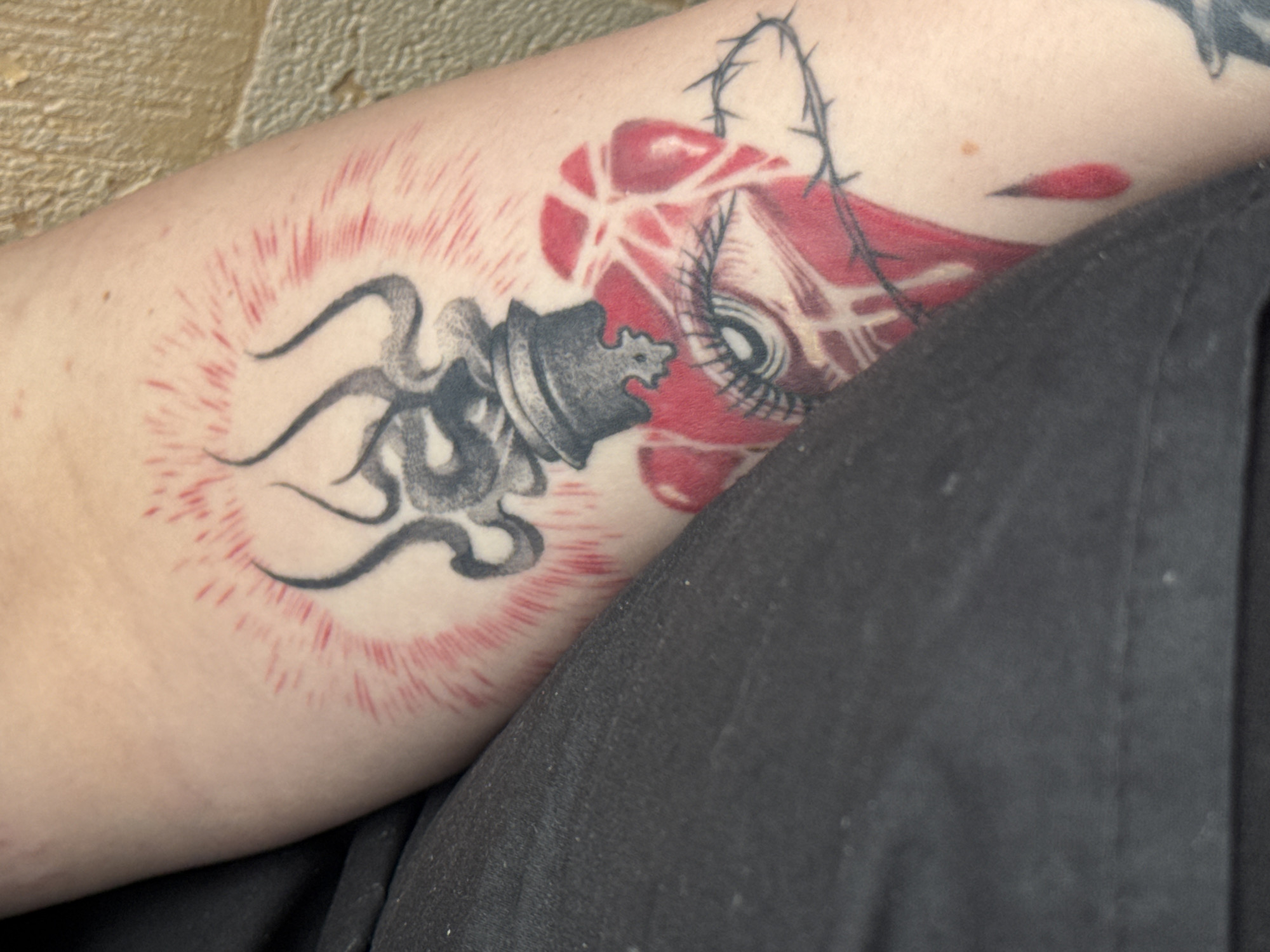 This media file displays a close-up view of a tattoo on a person's arm. The tattoo features a stylized representation of a chess piece, possibly a king, with black, swirling lines emanating from it, creating a dynamic, almost explosive effect. Surrounding this element is a radiating pattern of thin red lines. To the right of the chess piece, a red abstract shape is depicted, with fine black lines suggestive of eyelashes or hair. A thin black line resembling a thorn branch arches over this red shape. A small, red, tear-drop-like shape is also visible. The skin tone is fair. The background consists of a textured surface, possibly a wall or ground, with a blurred dark fabric in the foreground, obscuring part of the arm. The tattoo's imagery evokes a sense of intensity and possibly turmoil. There is no visible text in the media file.