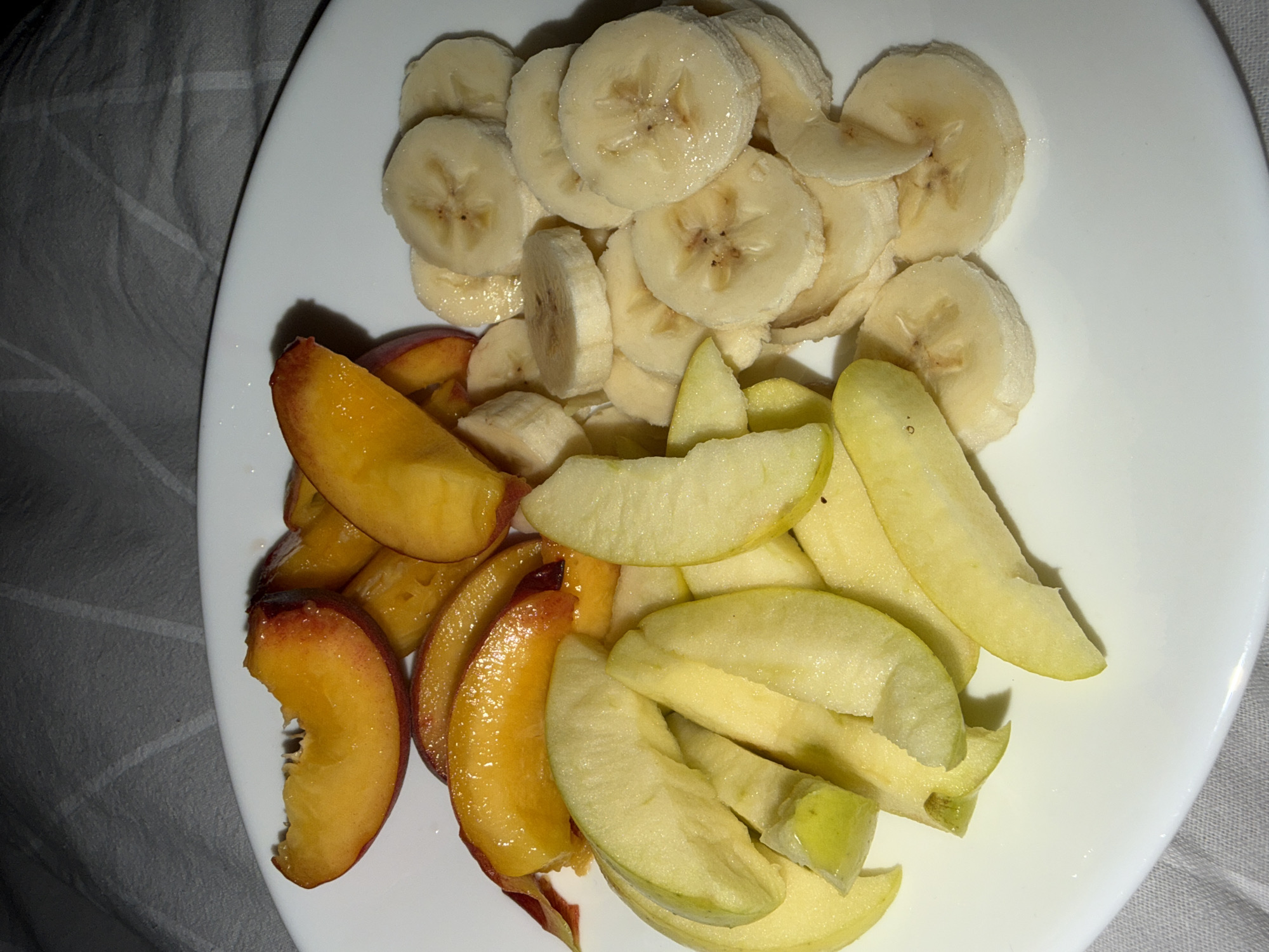 A white plate holds sliced fruit. On the left, there are wedges of apple, some appearing slightly bruised, with their skins still attached to some pieces. To the right of the apples are slices of banana, revealing their characteristic star-shaped cross-sections. In the upper left portion of the plate, there are slices of peach, their skin showing a gradient of red and yellow, and the flesh is a vibrant orange. The fruit is arranged on the plate without any discernible pattern, and the plate itself is resting on a textured, grey surface, likely a tablecloth or placemat. The background is dimly lit, suggesting an indoor setting. There is no information available to determine the location of this scene in Pazardzhik, Bulgaria.