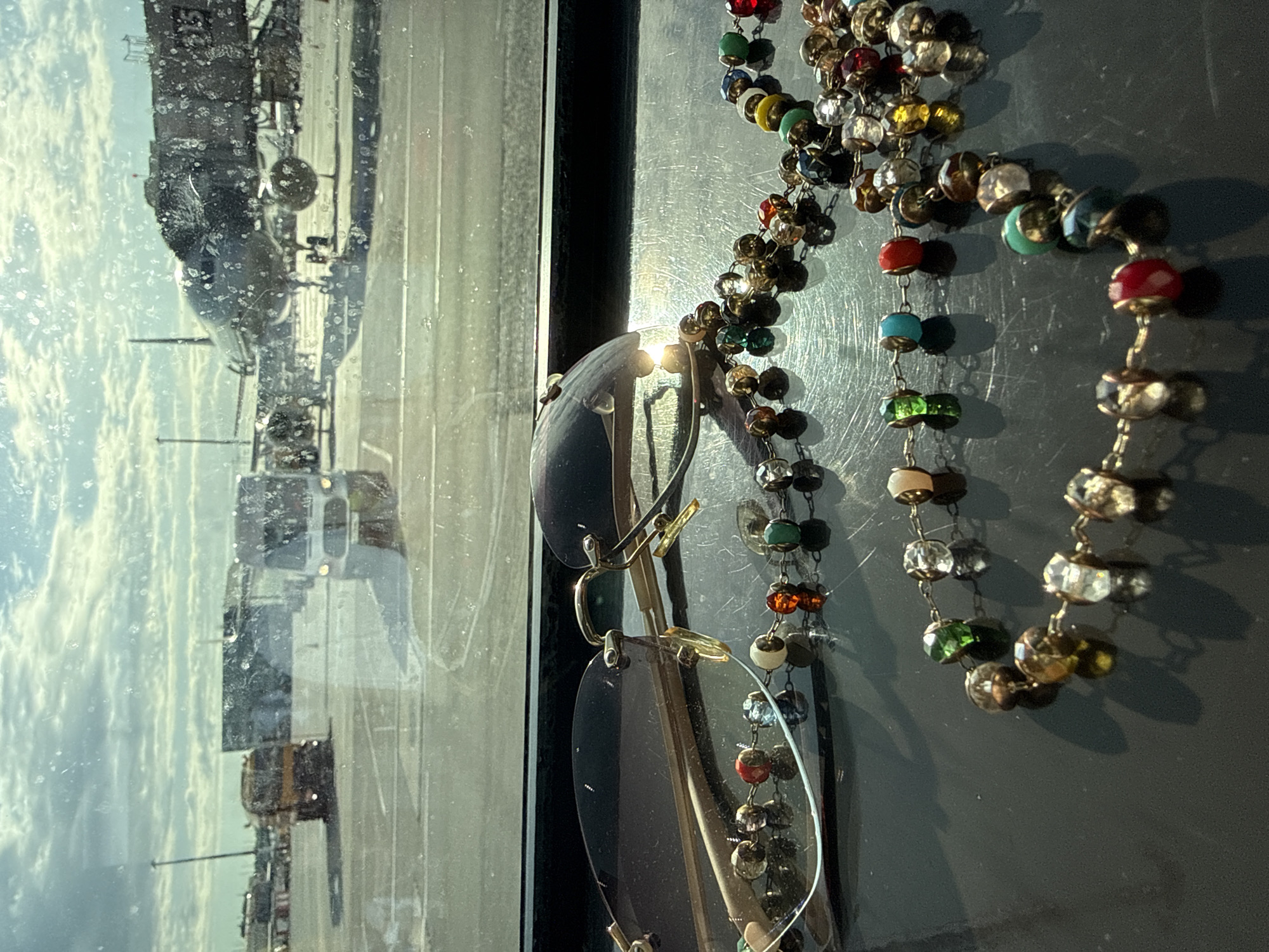 A photograph taken from an elevated position, likely indoors, shows a view through a window. The window reflects an outdoor scene of what appears to be an airfield or industrial area with various vehicles and aircraft visible in the distance, under a partly cloudy sky.
In the foreground, hanging near the window, are a pair of rimless eyeglasses and a strand of beaded jewelry. The beads vary in color and material, including some that appear to be glass and others that have a metallic sheen. The eyeglasses have gold frames and reflect the light coming in through the window.
No people are visible in the immediate frame. The image suggests a still life scene composed of personal items against the backdrop of an external environment. There is no definitive information about the location being in Khimki, Russia.