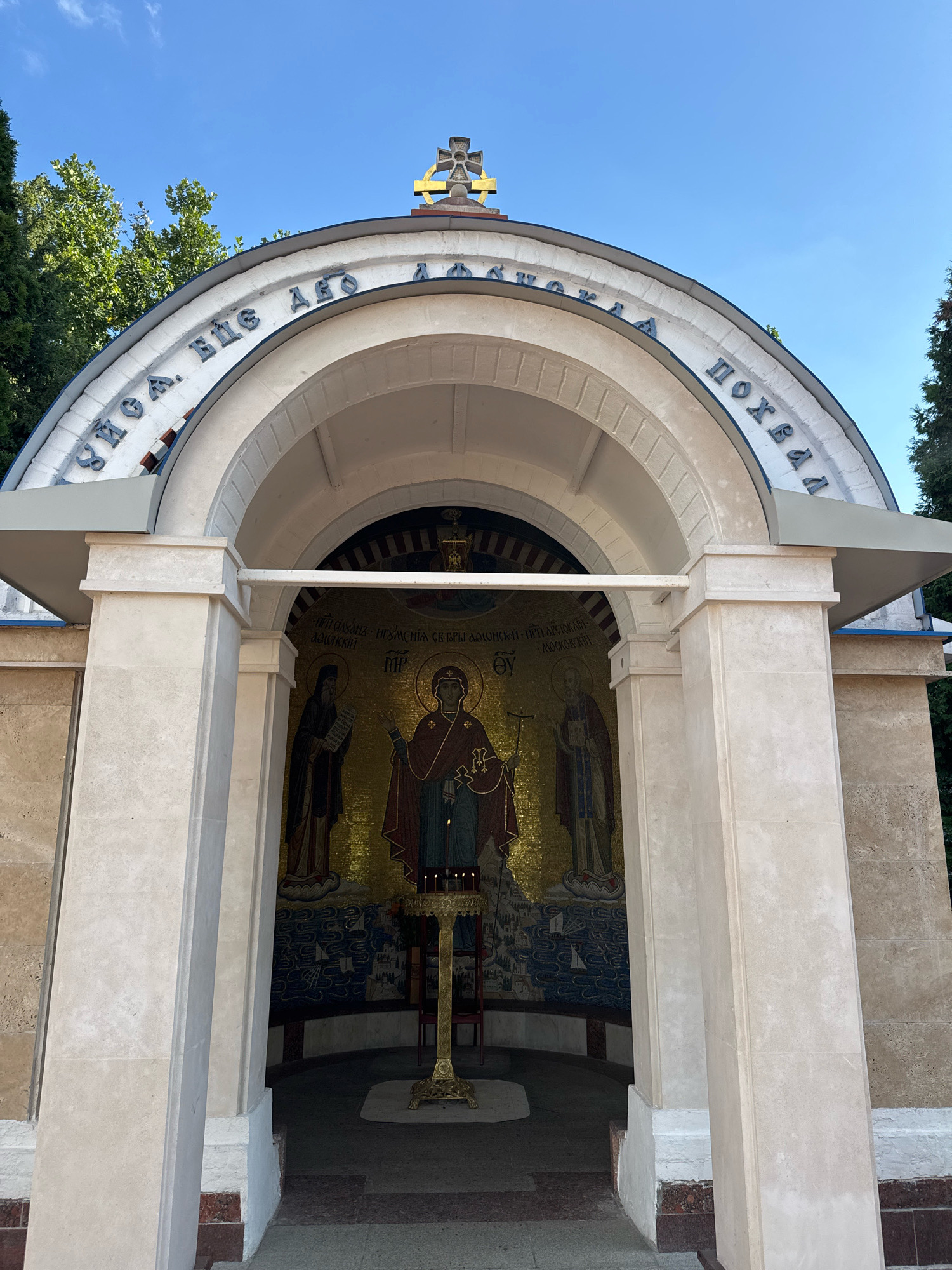 The image shows the facade of a small chapel in Moscow, Russia. The chapel has a semicircular archway supported by two stone pillars. Above the archway, there is an inscription in Cyrillic letters and a golden cross. Inside the chapel, a mosaic depicts religious figures, and a candle stand with lit candles is placed in front of it. The exterior walls are made of light-colored stone. Green trees are visible in the background, and the sky is clear and blue.