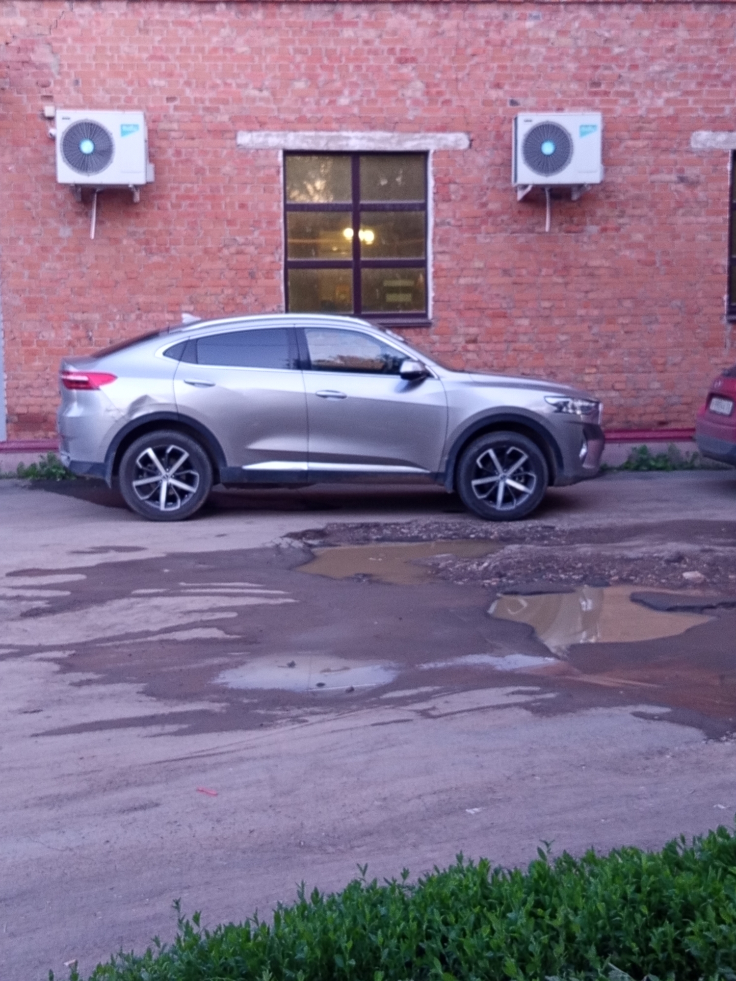 A silver car is parked outside a brick building in Novotroitsk, Russia. The car is a coupe-style SUV with tinted windows and black rims. In front of the car, the ground is muddy and uneven, with several puddles of water. The building has a red brick facade with white trim around a window and two air conditioning units mounted on the wall. One of the air conditioning units is on the left side of the window, and the other is on the right. The window is rectangular and appears to be a double-paned window with a dark frame. The building is located on a street, as evidenced by the presence of the car and the road surface. The sky is overcast, and the lighting suggests it is either early morning or late evening. The overall scene is one of an urban environment, possibly a residential or commercial area.