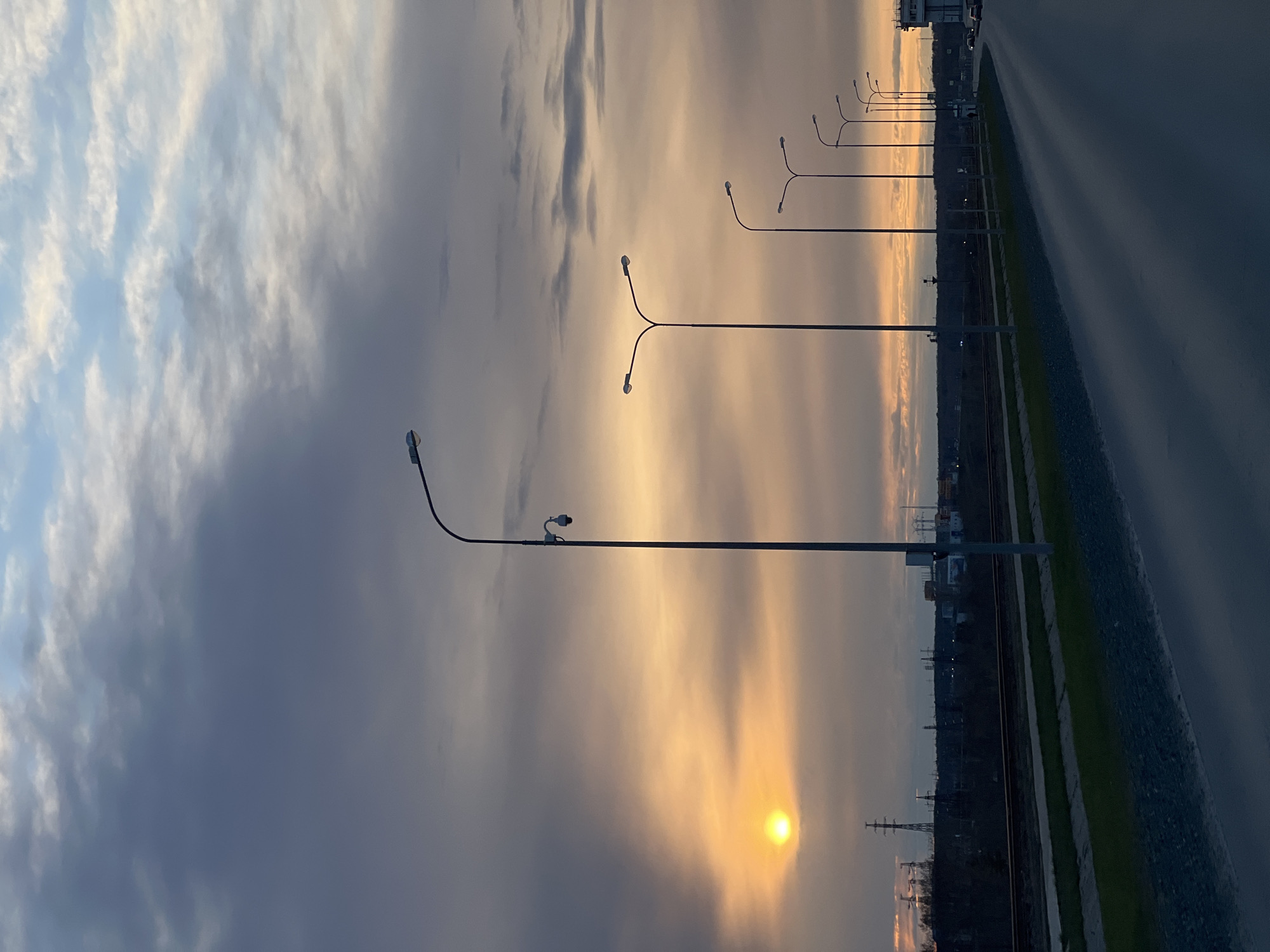 The image shows a landscape at dusk in Chaikovsky, Russia. A row of streetlights stretches along a road. The sky is filled with clouds, illuminated by the setting sun. The sun's light creates a golden glow in the sky and reflects on the road surface. A railway track runs parallel to the road, with vegetation between them. Buildings are visible in the background. The lighting conditions suggest twilight.