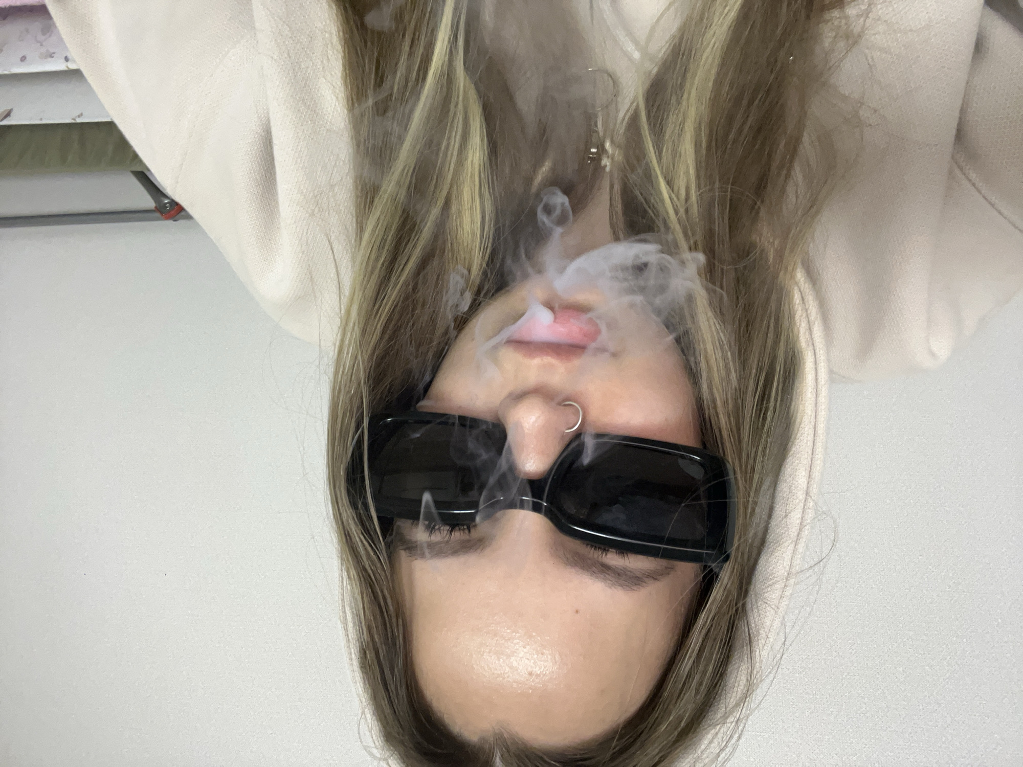 The originally inverted image depicts a close-up, upright view of a young person, likely female, with fair skin and long, straight, light brown hair featuring blonde highlights. She is wearing dark, rectangular sunglasses that obscure her eyes, and a silver hoop nose ring is visible on her right nostril. Her lips are slightly parted, and a prominent cloud of white smoke or vapor emanates from and around her mouth and nose, rising upwards.
She is dressed in a casual, light-colored, possibly beige or off-white, hooded sweatshirt or top. A small, simple earring is visible on her right ear, partly concealed by her hair. The background consists primarily of a plain, light-colored wall, suggesting an indoor setting. In the upper left corner of the original image (which would be the lower right when viewed upright), a small section of a shelf or cluttered surface with a red clip or object is faintly visible. The activity taking place is the exhalation or blowing of smoke/vapor. No specific time of day or weather is discernible. The overall mood conveyed is casual and somewhat mysterious due to the sunglasses and smoke. No visible text is present in the image.