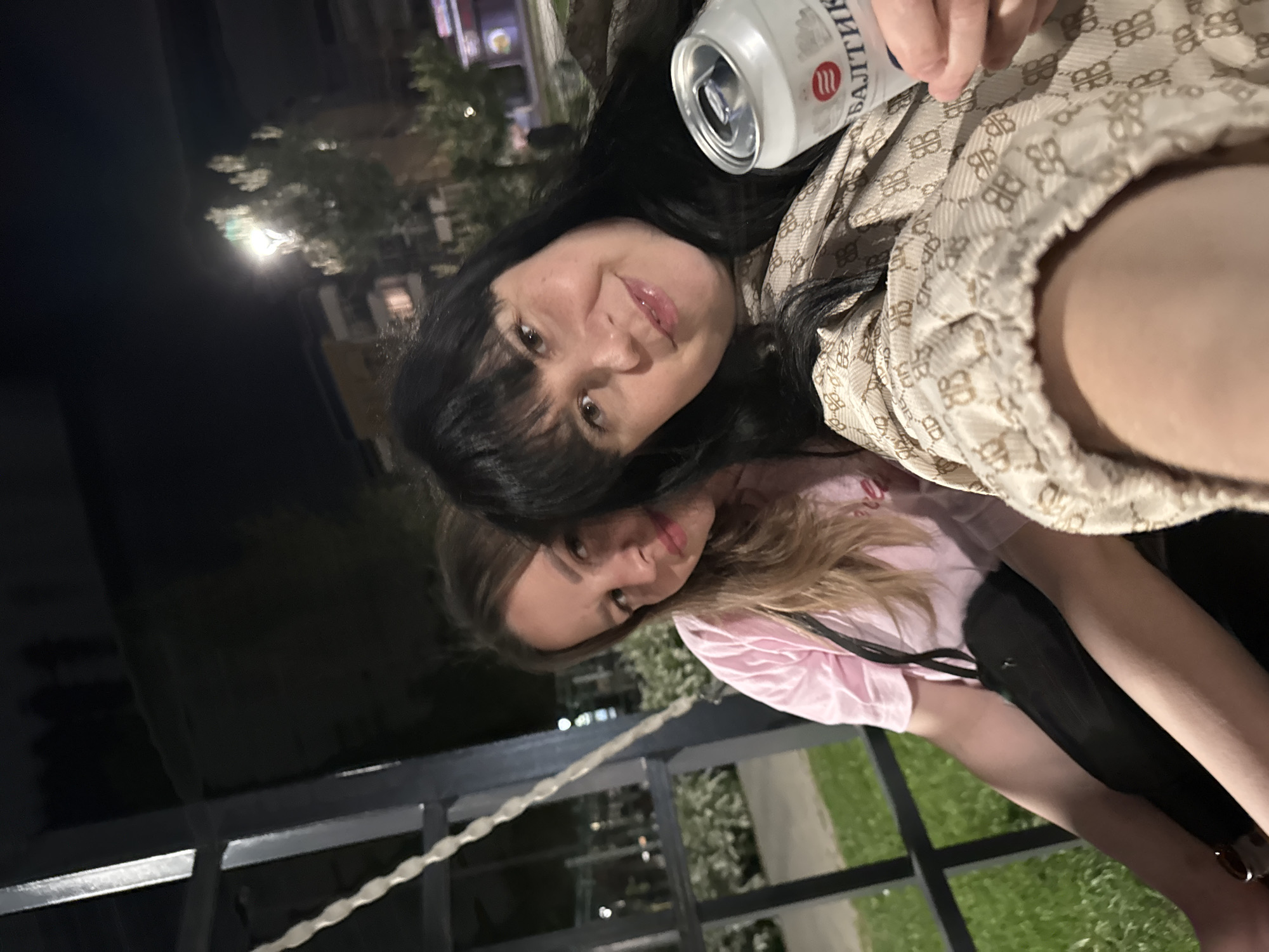 Two women are captured in a selfie. The first woman has dark hair, and she's holding a can of Baltika beer. The second woman has blonde hair. They appear to be outdoors, possibly at night, given the lighting. A metal railing and greenery are visible in the background. The location is Zav'yalovo, Russia.