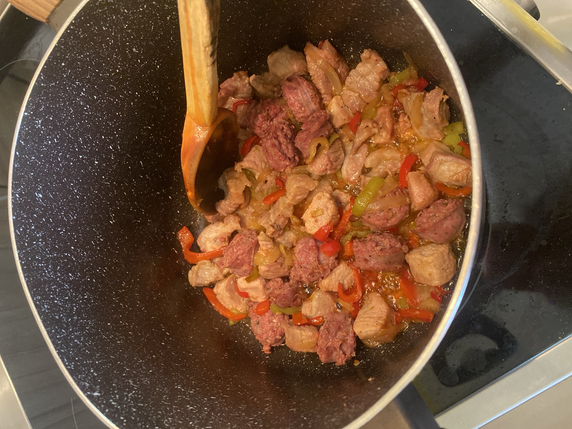 A high-angle, close-up shot captures a meal being cooked in a large, dark non-stick pot on a stovetop. The pot, featuring a speckled black and white interior, is filled with a mixture of various ingredients simmering in a glossy, reddish-orange liquid.
The main components of the dish include numerous chunks of light-colored meat, likely pork or chicken, which appear partially cooked or browned. Interspersed among the meat are generous slices of reddish-pink sausage with a coarse, crumbly texture. Brightly colored vegetables are also visible, specifically chopped red and green bell peppers, along with translucent slices of onion.
A wooden spoon with a reddish-brown handle is partially submerged into the food, resting along the rim of the pot, suggesting recent stirring or readiness for it. The immediate surroundings show a black electric stovetop surface, with reflections indicating a smooth finish. The bright edge of the pot's rim is also visible. The scene depicts an ongoing culinary process in a kitchen setting. There are no people visible, no discernible text, and no cues to indicate the time of day or weather. While external information suggests the location is Sliven, Bulgaria, this is not visually evident in the image itself.