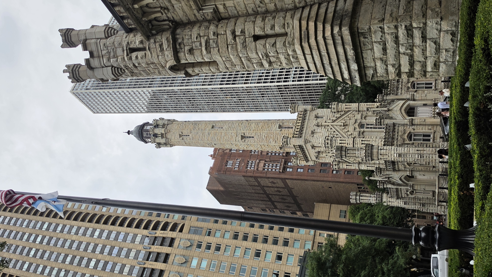 This is a low-angle, exterior, daylight photograph of the Chicago Water Tower and surrounding buildings. The Water Tower is a Gothic Revival style building constructed of limestone and located at 806 North Michigan Avenue, Chicago, Illinois, United States of America. The tower is flanked by a tall, light-colored modern skyscraper to its right and a taller, darker brick building with patterned masonry to its left. In the foreground, there is a well-manicured hedge with several downlights. A black lamppost stands prominently to the left of the frame. Several people are visible in front of the Water Tower and on the sidewalk. An American flag and a Chicago flag are flying from the lamppost. The sky is overcast.