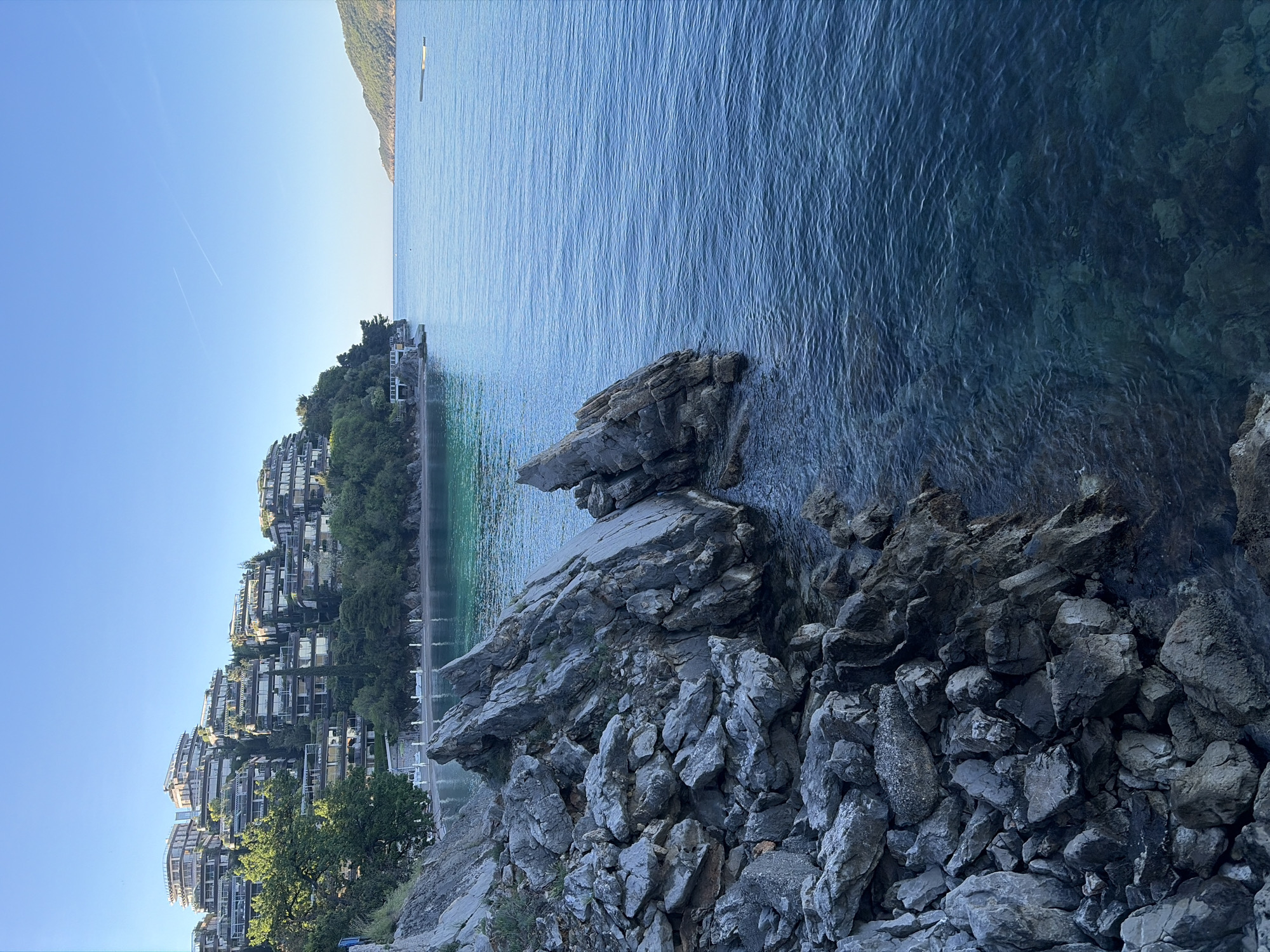 The image captures a coastal scene in Budva, Montenegro. In the foreground, a rocky terrain slopes down towards the Adriatic Sea. The water is clear and ranges in color from light turquoise to deep blue. To the left, a series of modern apartment buildings rise up a hillside, interspersed with trees. The sky is clear and blue. In the distance, a narrow strip of land or island is visible. There are no people depicted in the scene.