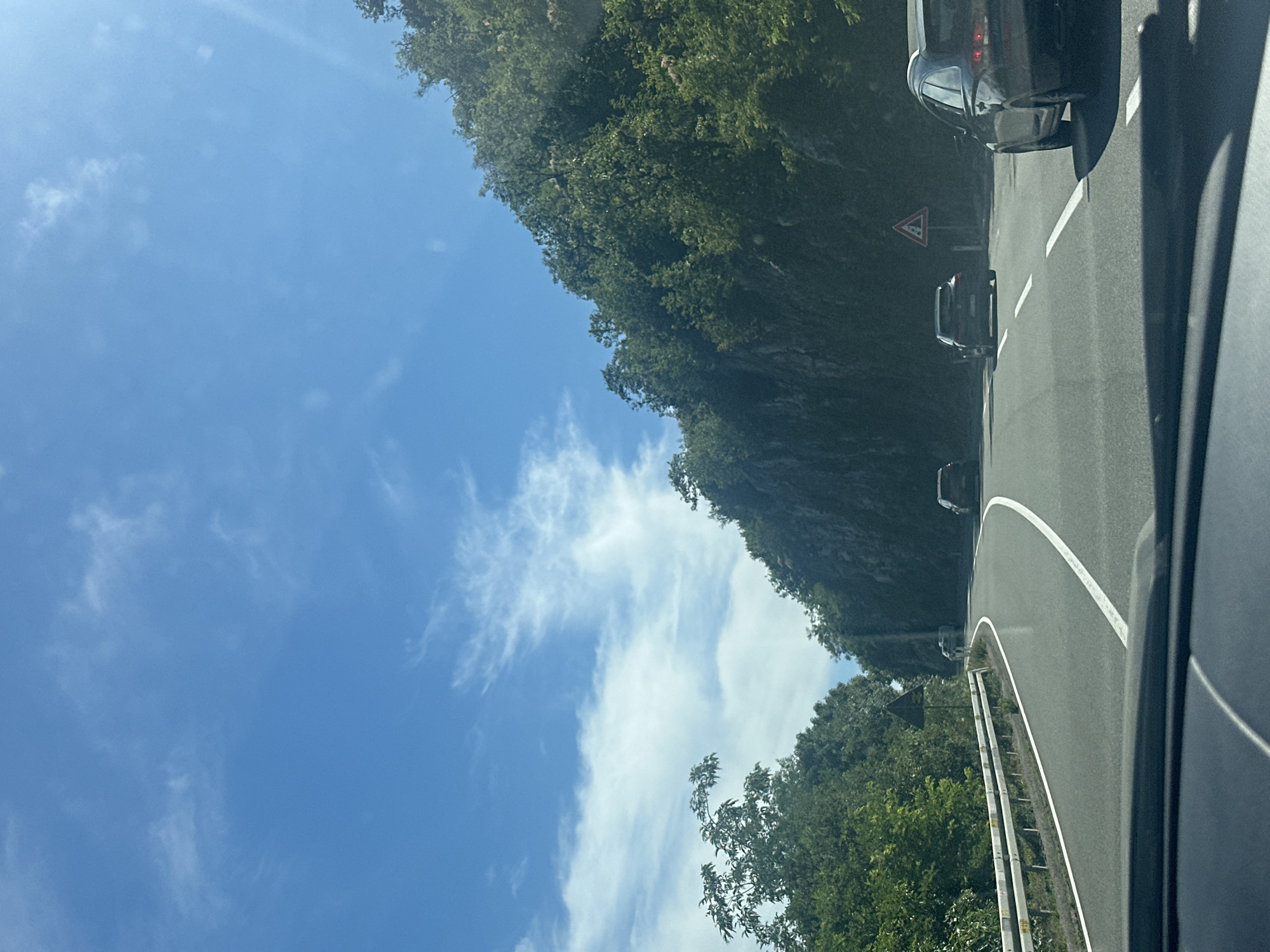 The image depicts a view from inside a vehicle driving on a winding, paved road in Donji Ulići, Montenegro. The road has clear white lane markings. On the left side of the road, a white guardrail runs along the edge, separating the road from a steep, tree-covered embankment. The right side of the road is bordered by a rocky cliff face densely covered with green foliage. Several cars are visible on the road: one dark-colored station wagon in the foreground right, another dark car further ahead, and a third car in the distance to the left. A triangular warning sign with a red border and a symbol indicating falling rocks is posted on a pole to the right of the road. The sky is a bright blue with scattered white clouds. The sun is shining, casting shadows on the road and vegetation. The scene appears to be a rural or mountainous area.
